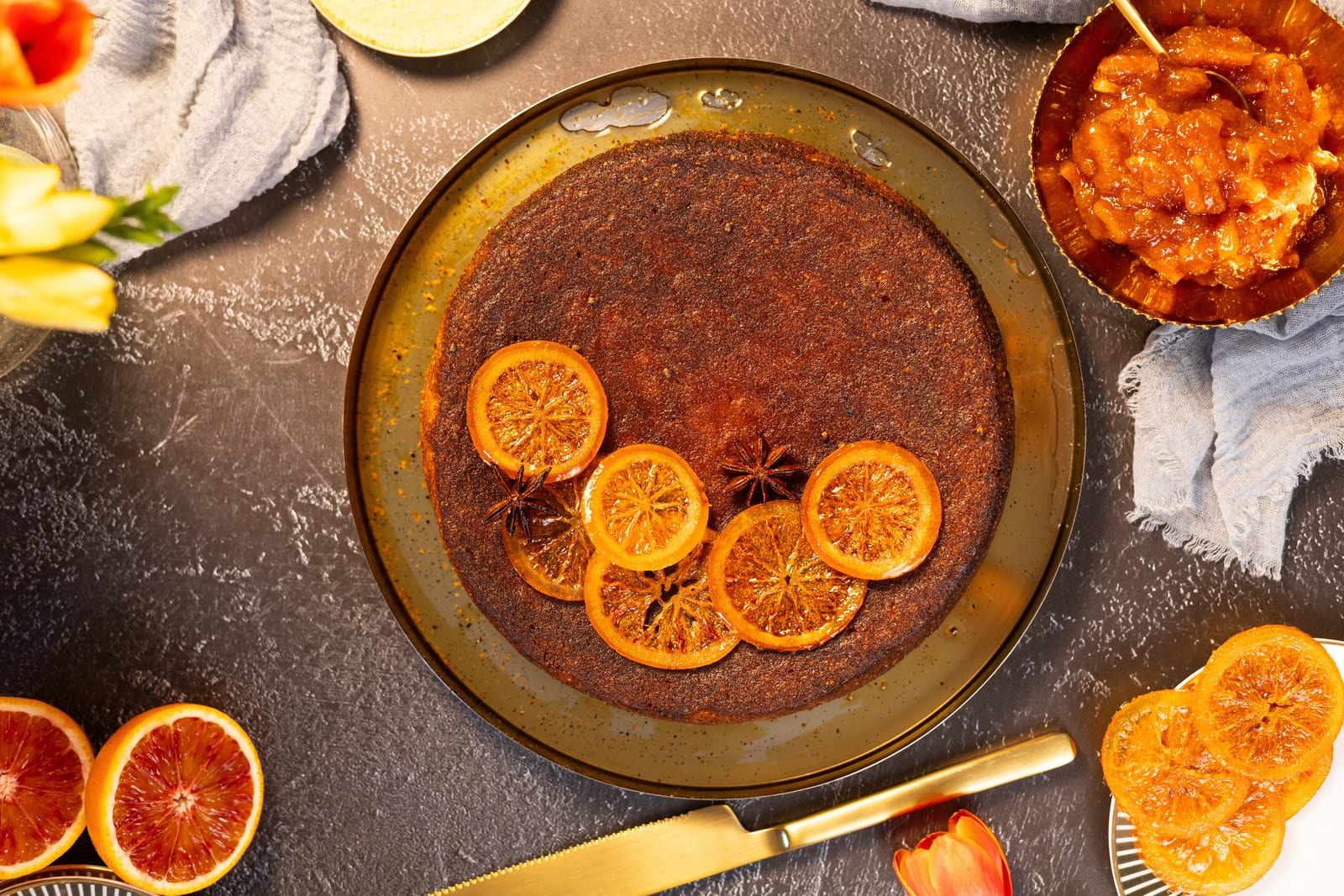 Blood orange, almond and polenta cake from above, topped with candied orange slices and star anise, beside halved blood oranges