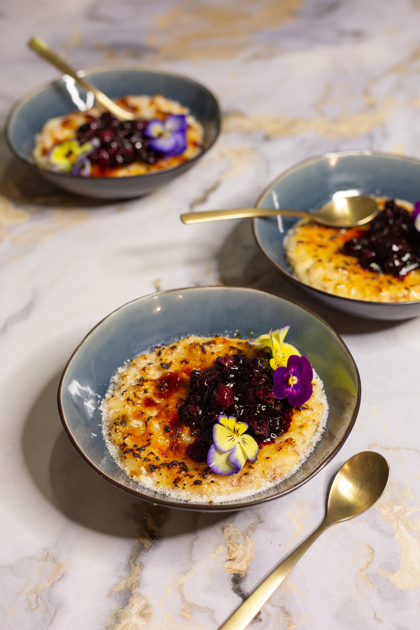 Three bowls of kheer brûlée with blueberry compote and violas on a marble surface