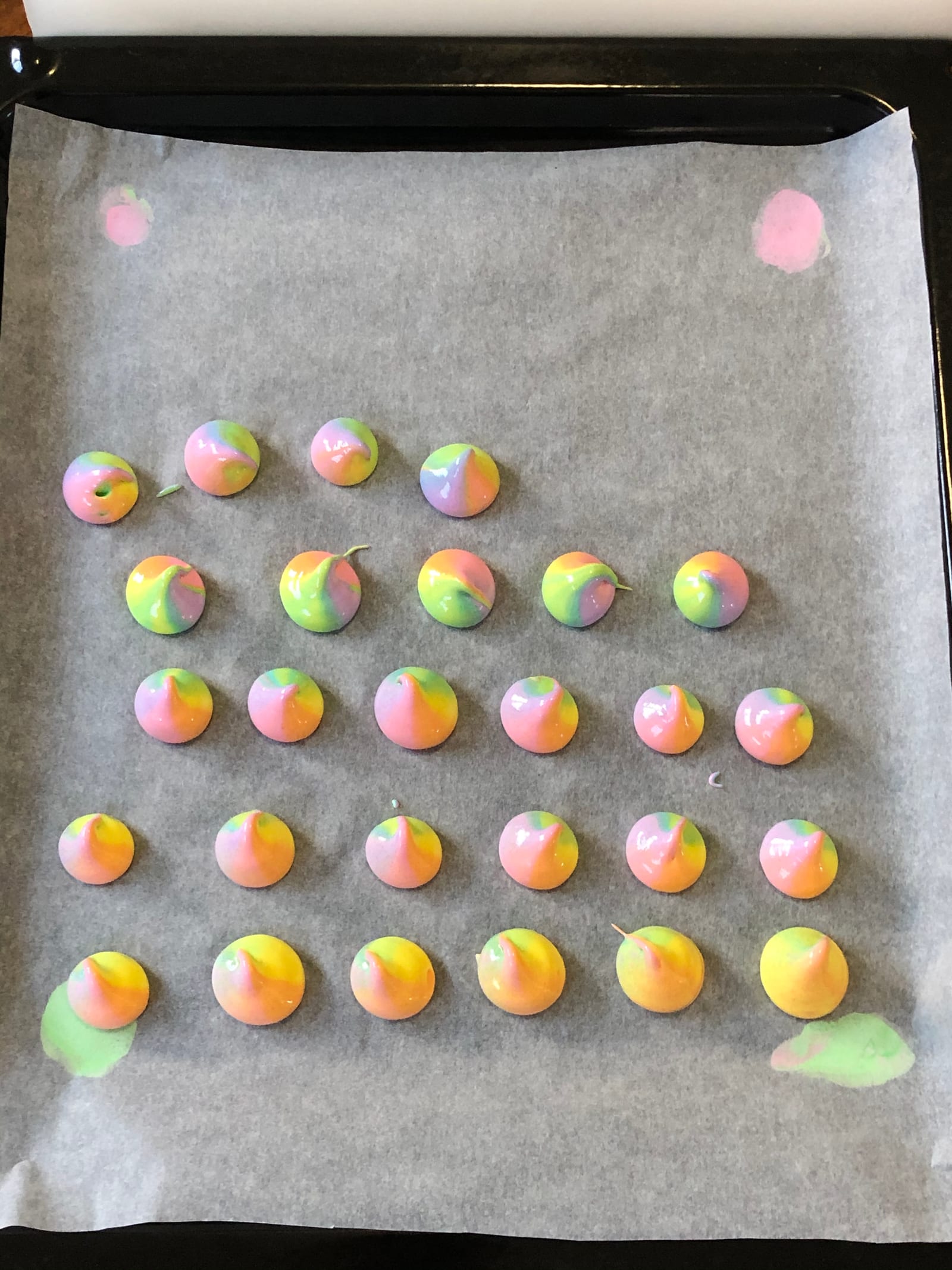 A baking tray of piped rainbow meringue kisses on parchment, from above