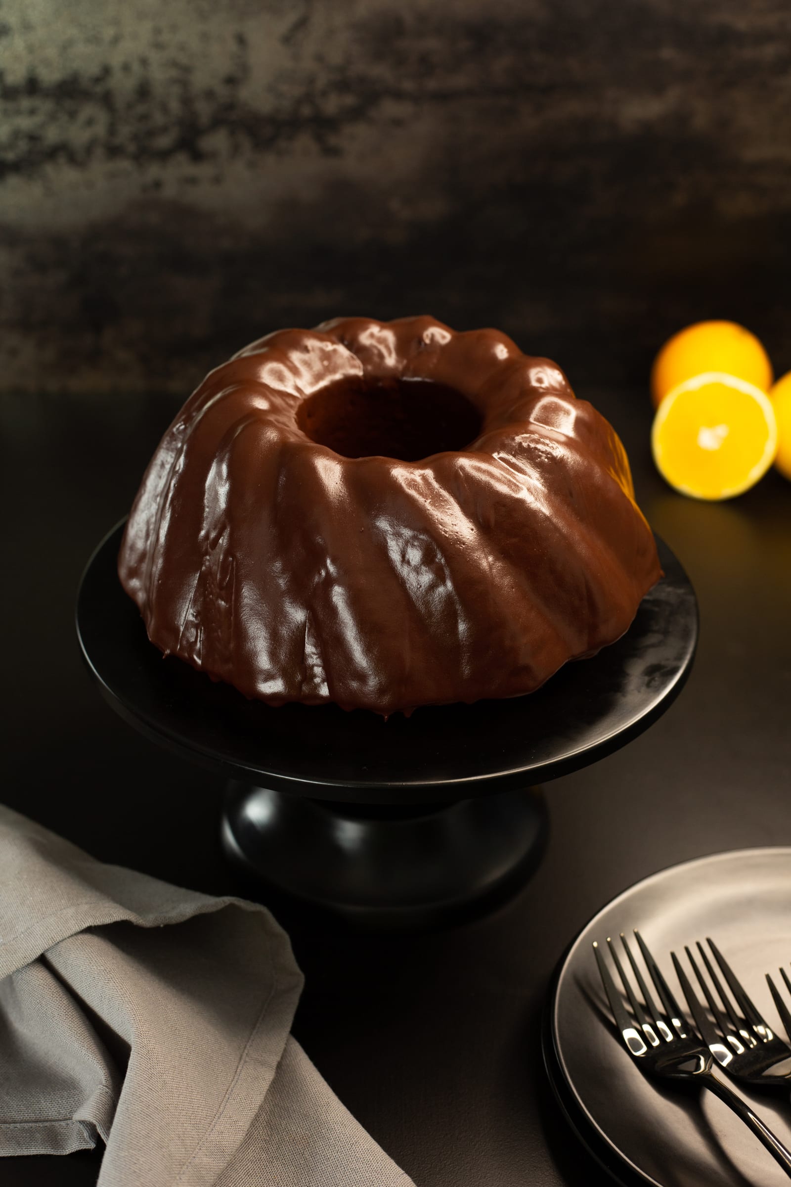 A whole orange and olive oil bundt in a dark chocolate glaze on a cake stand, with oranges and forks