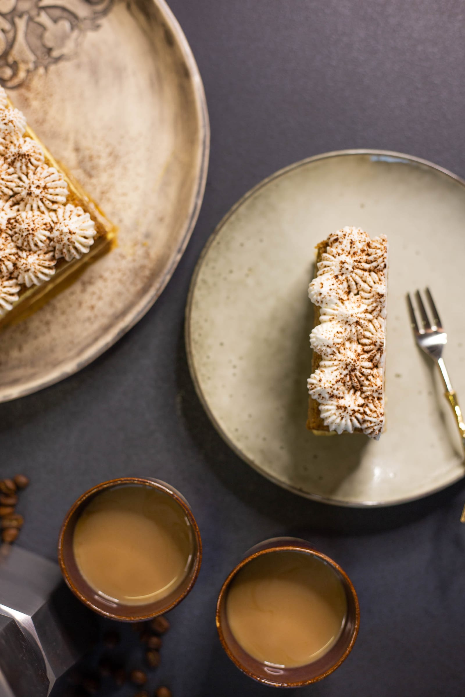A slice of tiramisu tres leches cake on a plate with cups of coffee