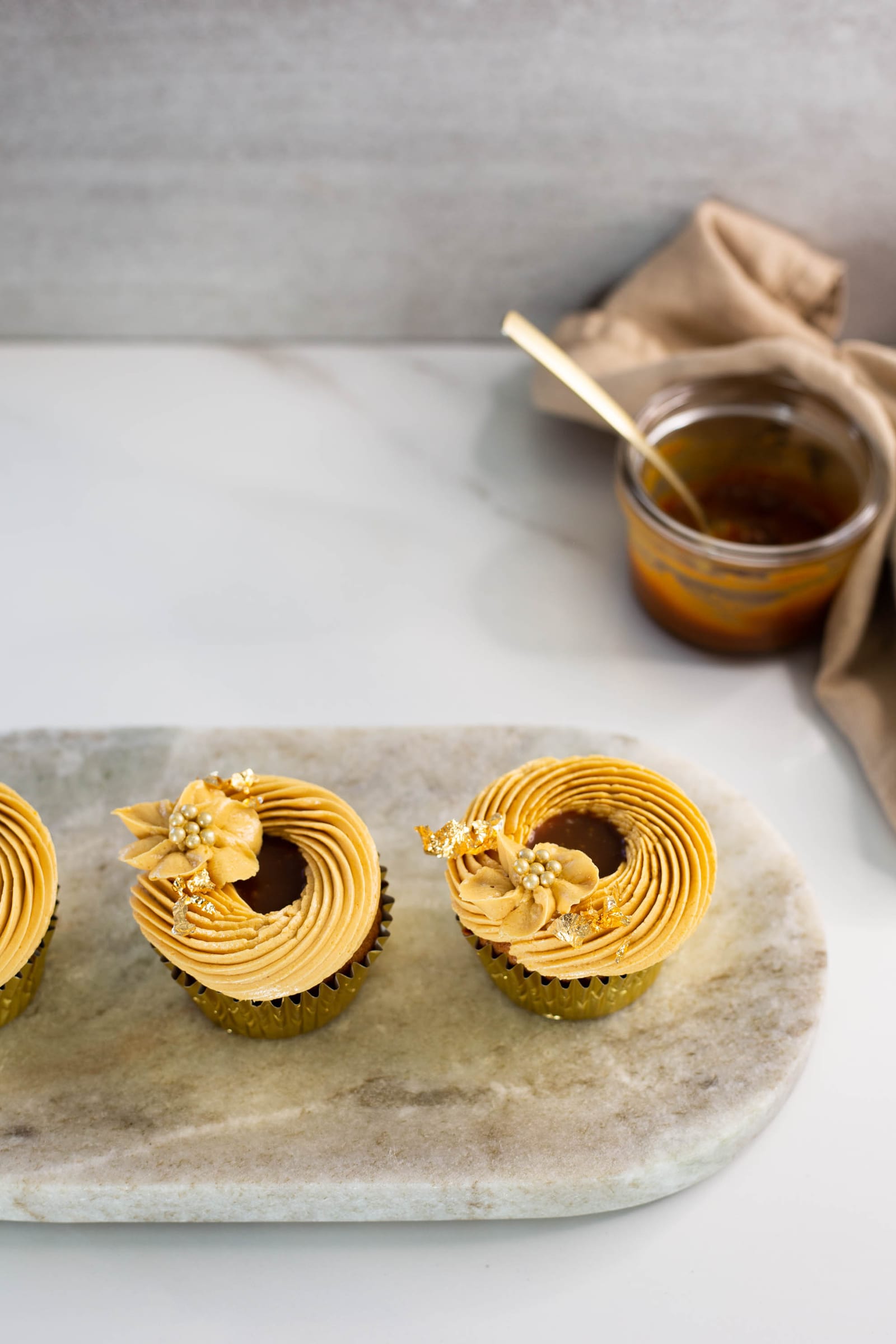 Three salted caramel cupcakes on a marble board beside a jar of salted caramel sauce
