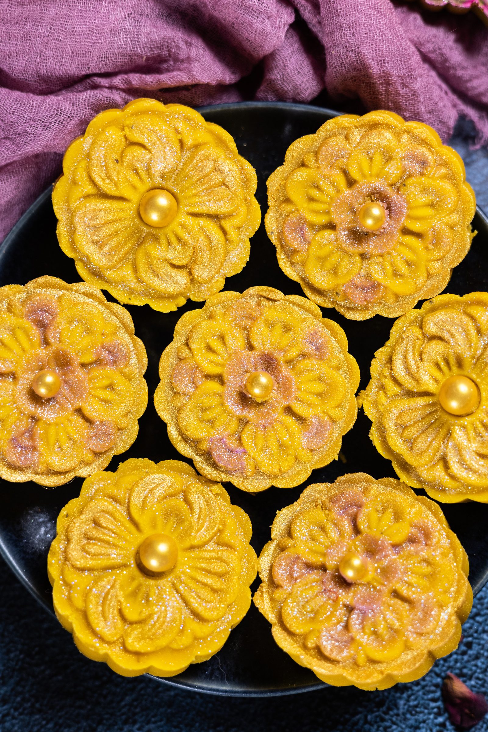Flower-shaped mango burfee topped with gold pearls on a dark plate, from above