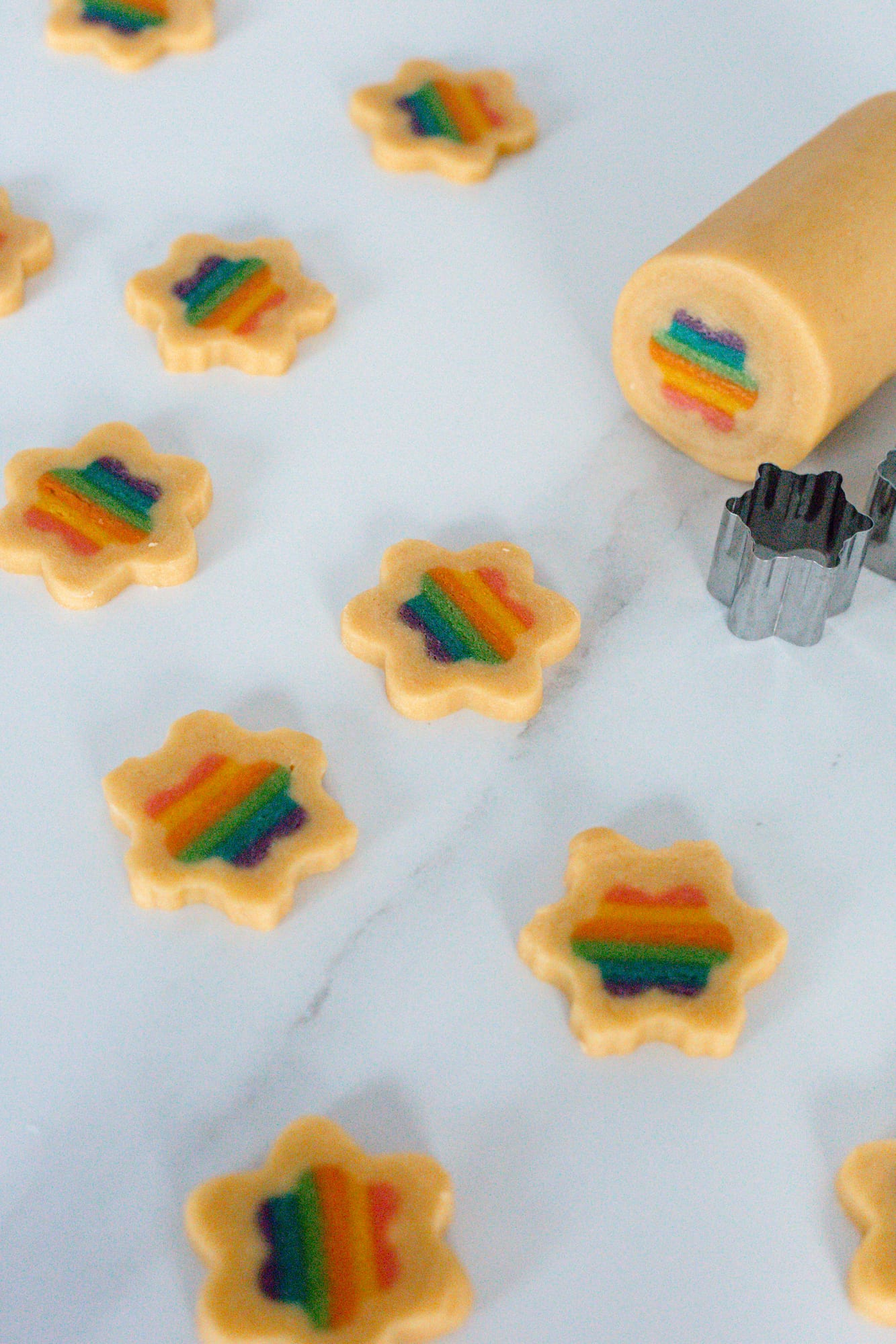 The log of plain dough with its rainbow centre showing at the cut end, a flower cutter and baked rainbow cookies