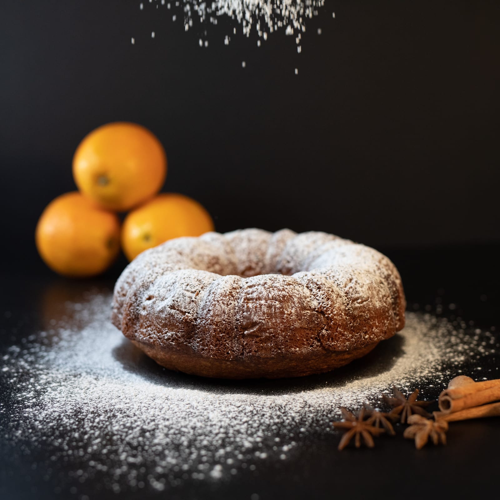 An orange spice pound cake dusted with icing sugar, with oranges and star anise