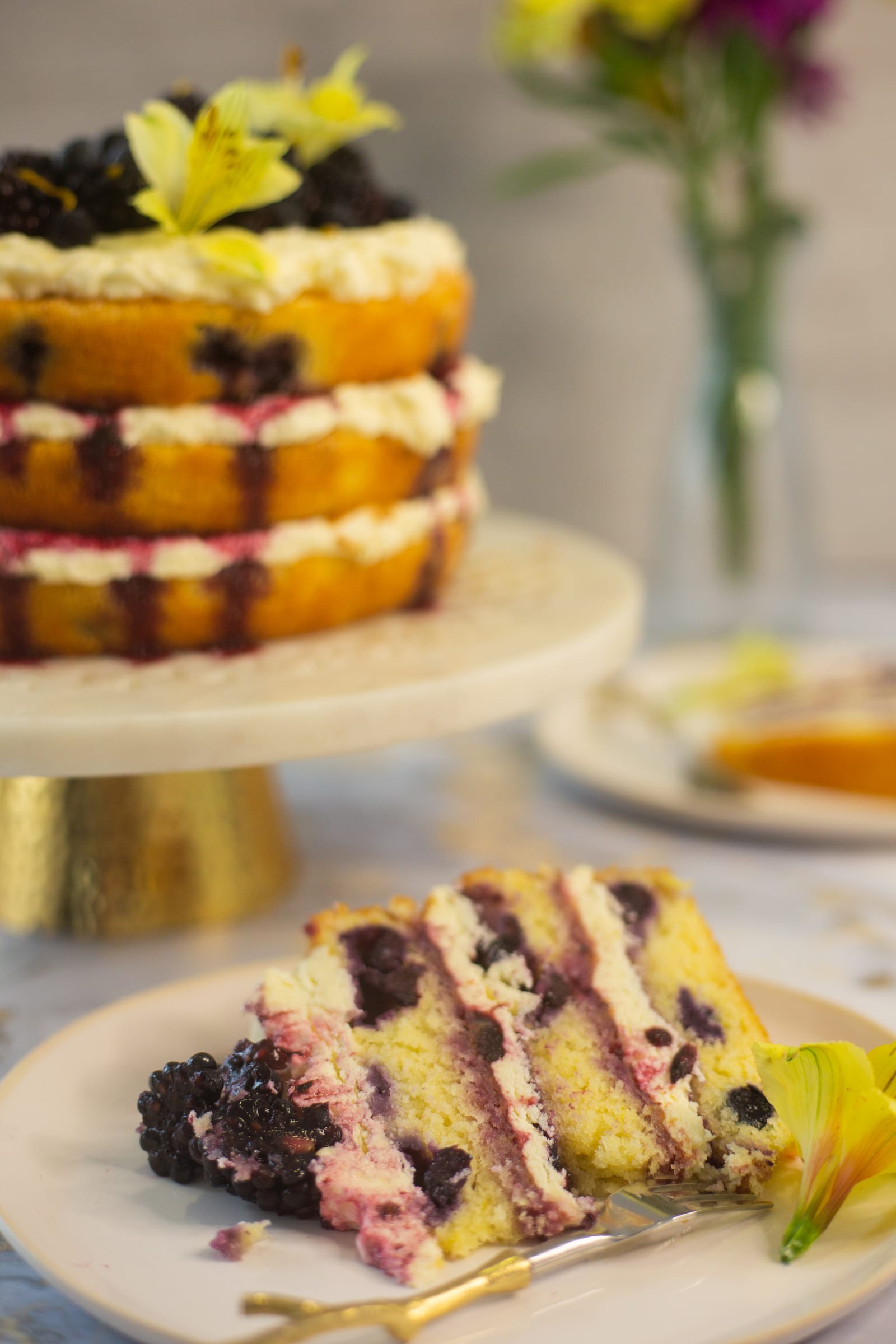 A slice of lemon and blueberry cake on a plate with the cut cake behind