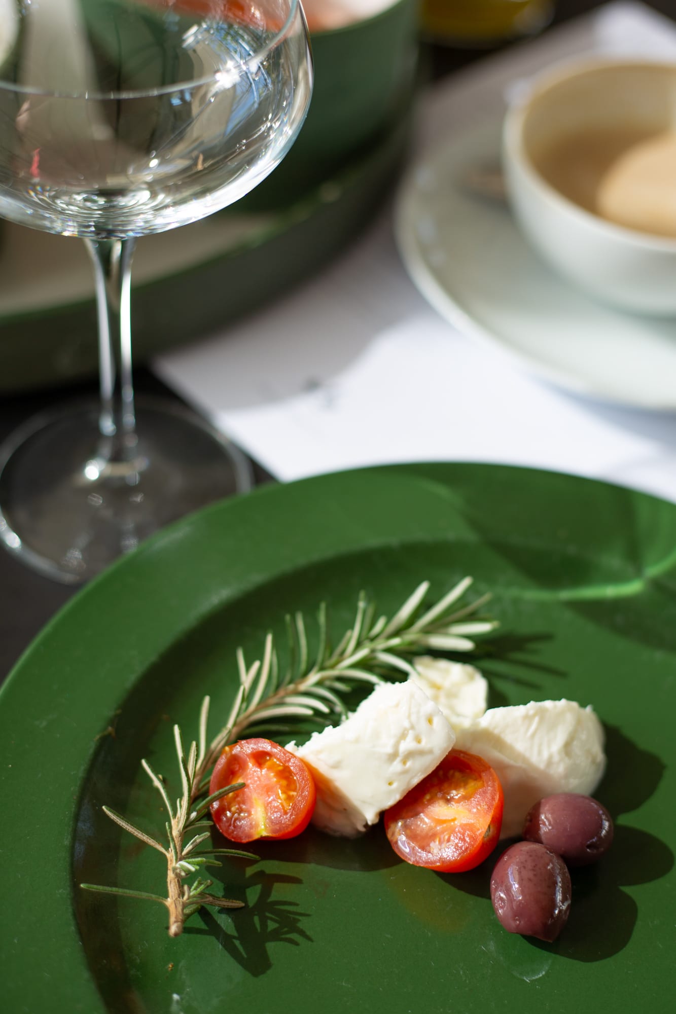 Mozzarella, halved cherry tomatoes, olives and a rosemary sprig on a dark green plate, with a wine glass and cappuccino behind