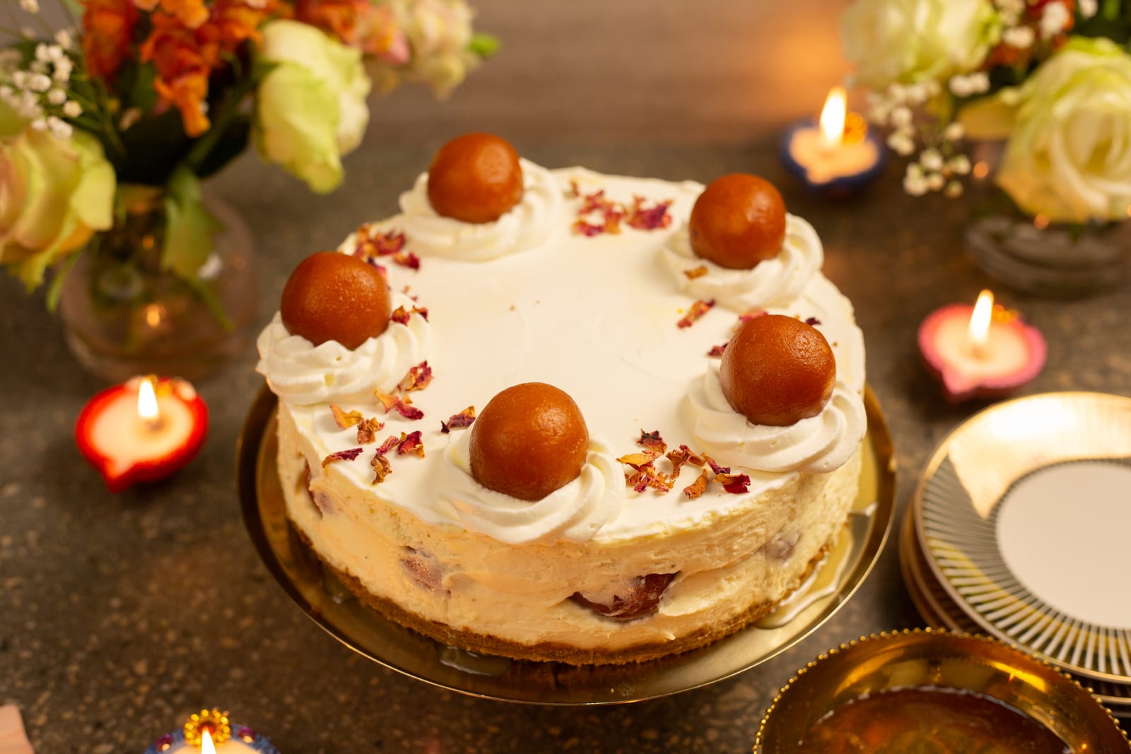 A gulab jamun cheesecake topped with cream rosettes and gulab jamun, with flowers and diyas