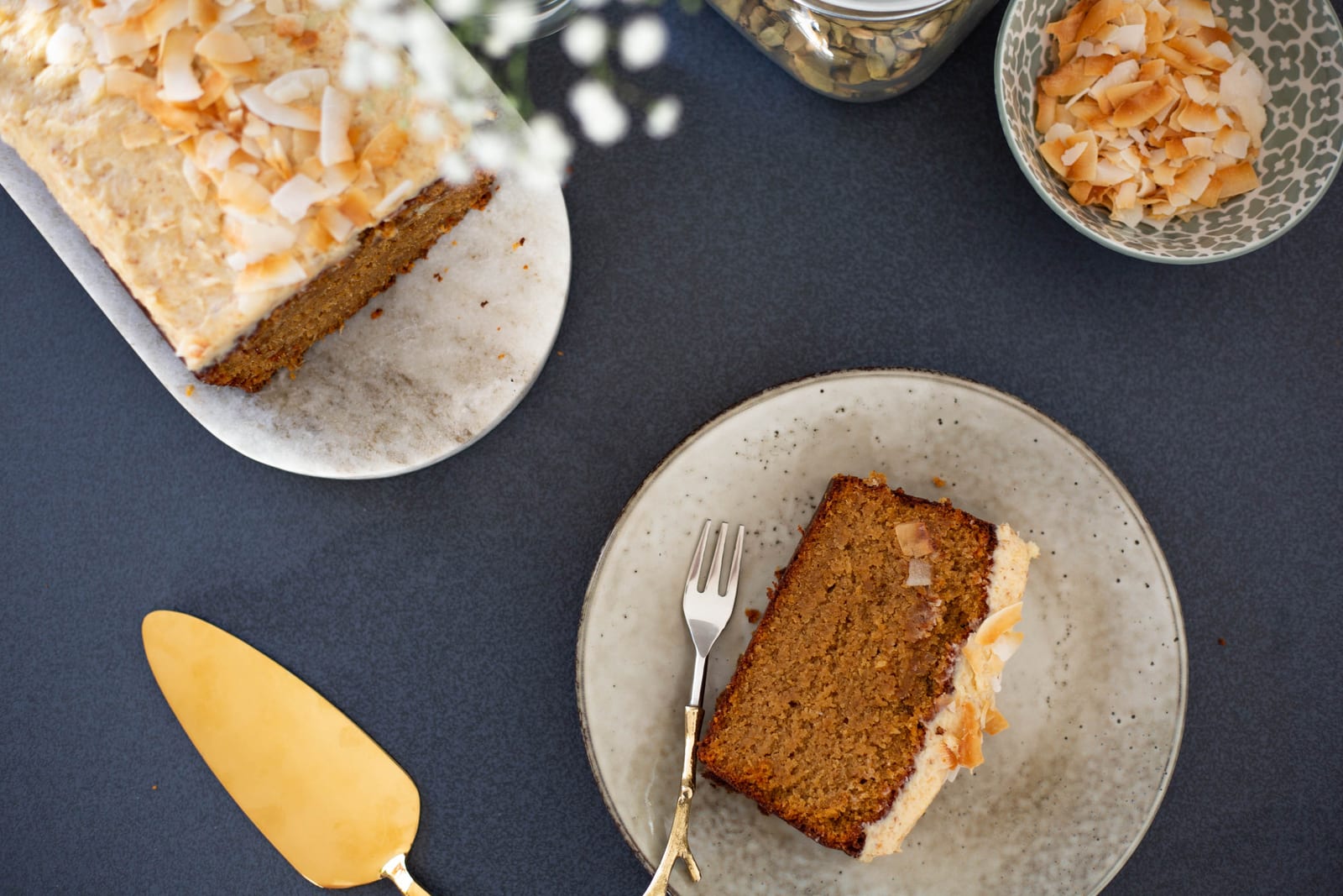 A slice of gluten-free coconut loaf on a grey plate with a fork beside the loaf and a bowl of coconut, from above