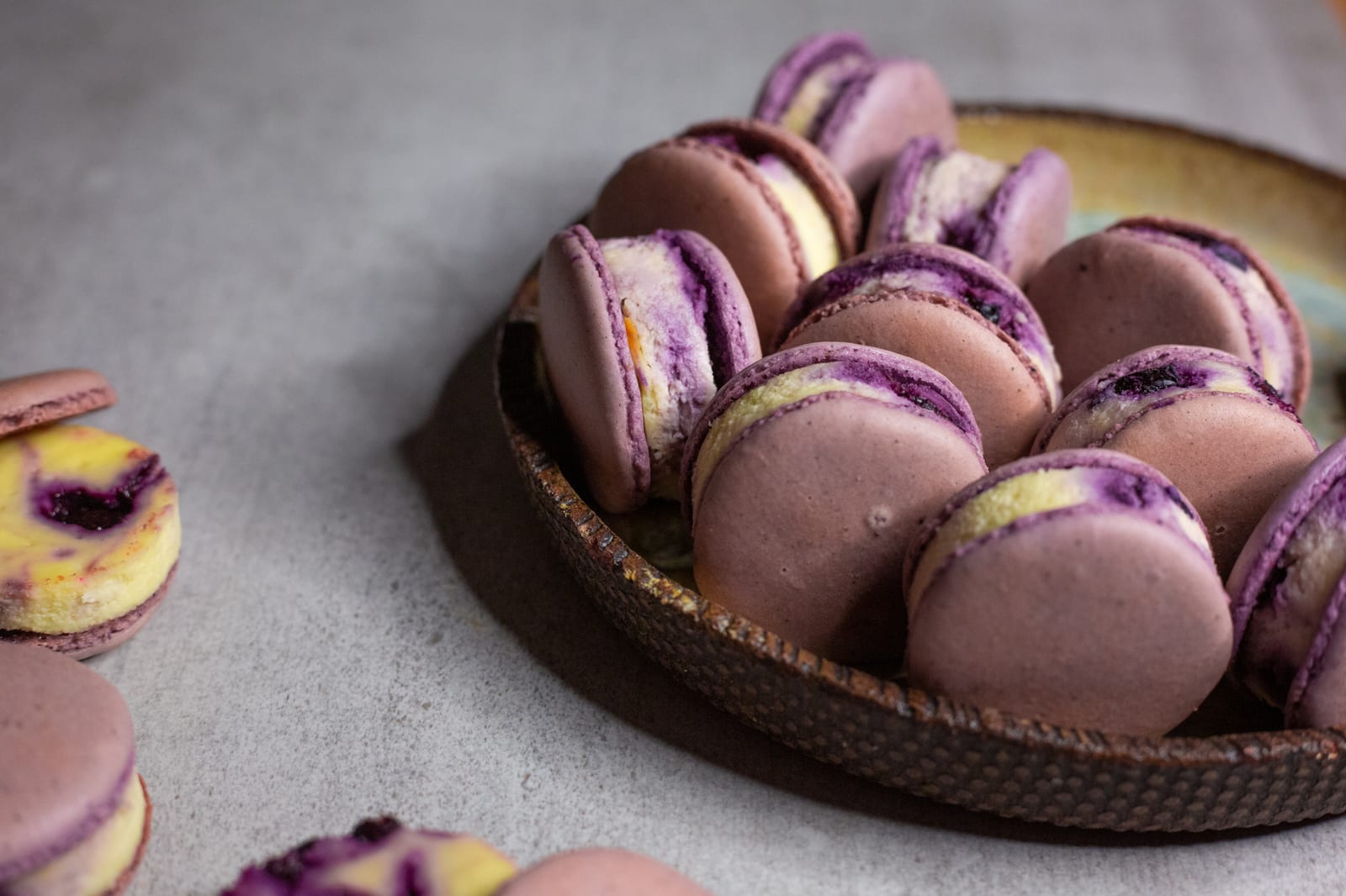 A plate piled with purple blueberry cheesecake macarons, one in the foreground