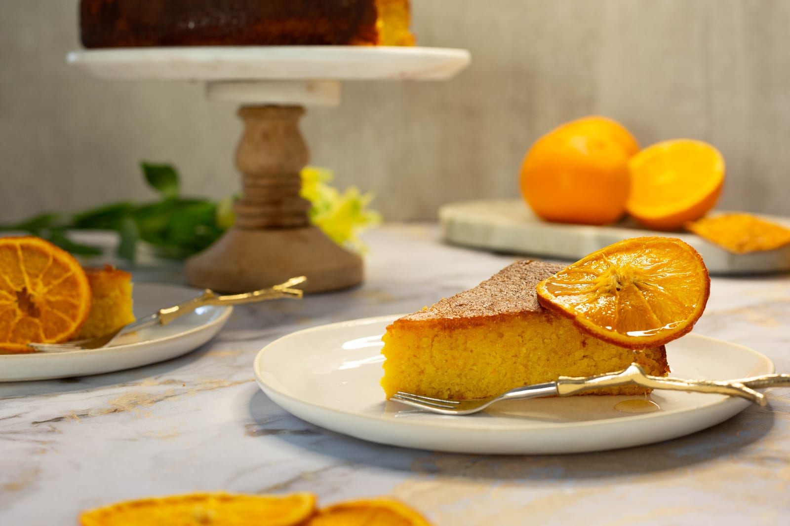 A slice of whole orange, cardamom and polenta cake in front of the cake on its stand, dried orange slices in front