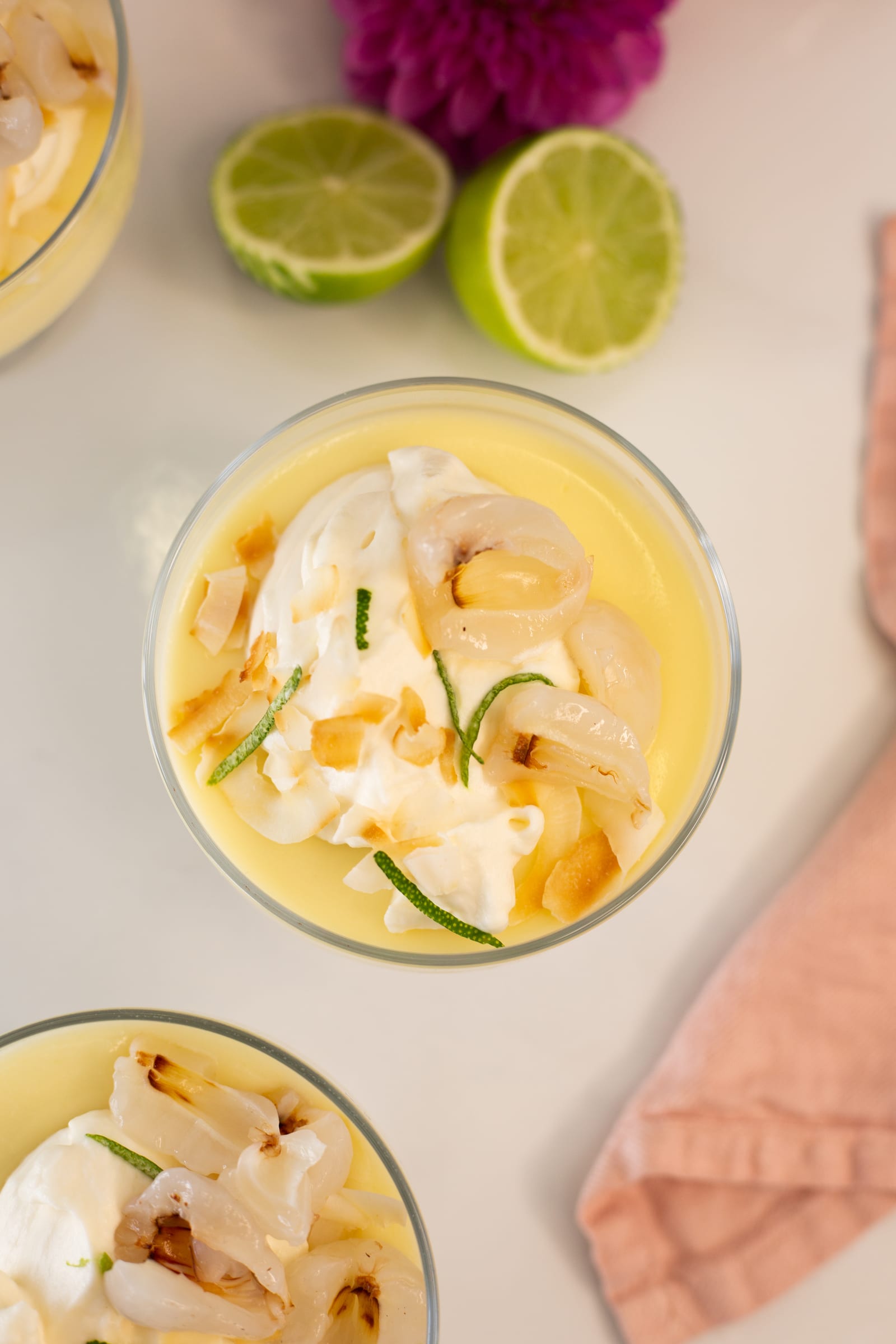 Looking down on a lime and coconut posset topped with whipped cream, litchi, toasted coconut and lime zest