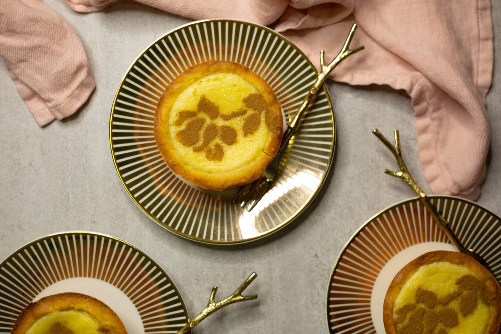 A mini milktart with a cinnamon rose stencil on a striped gold-rimmed plate beside a pink napkin, from above