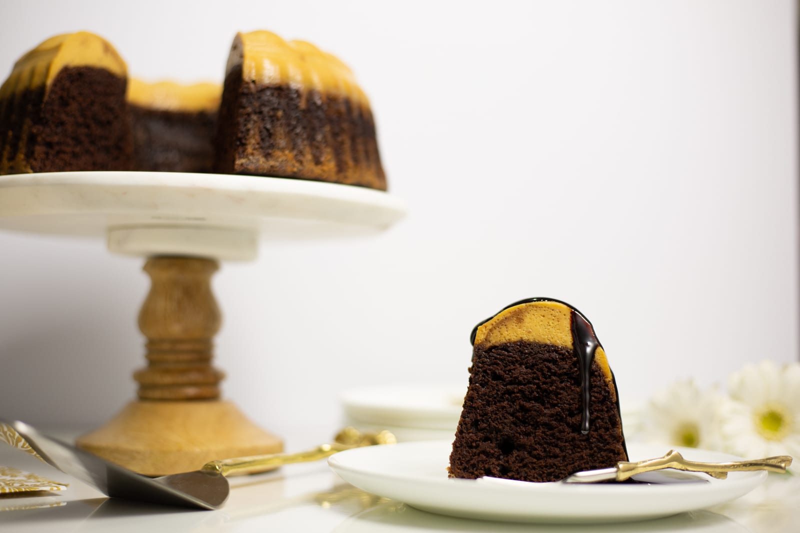 A cut caramel white chocoflan on its stand with a slice and chocolate sauce in front