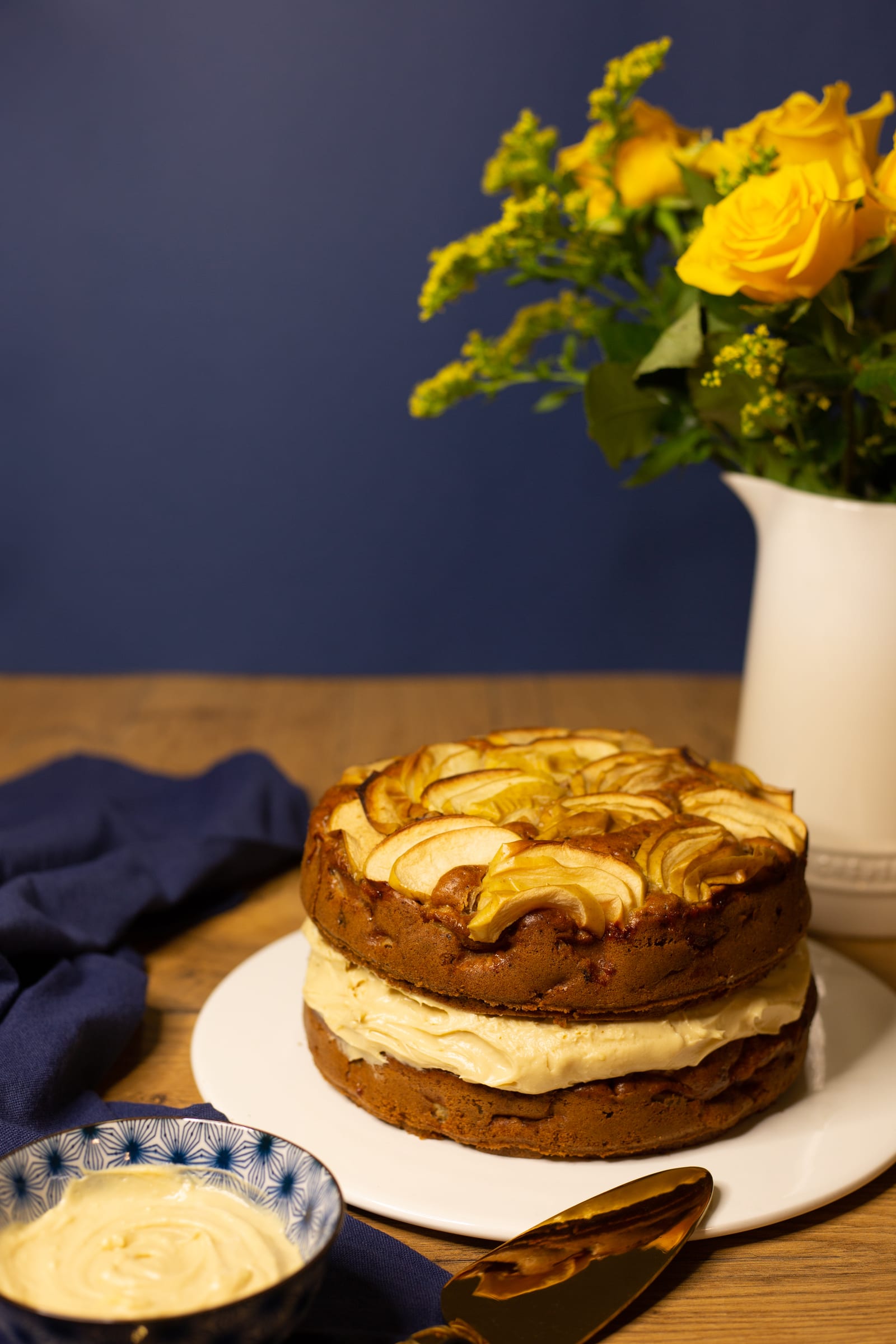 A two-layer apple and olive oil cake topped with sliced apples, yellow roses in a jug behind