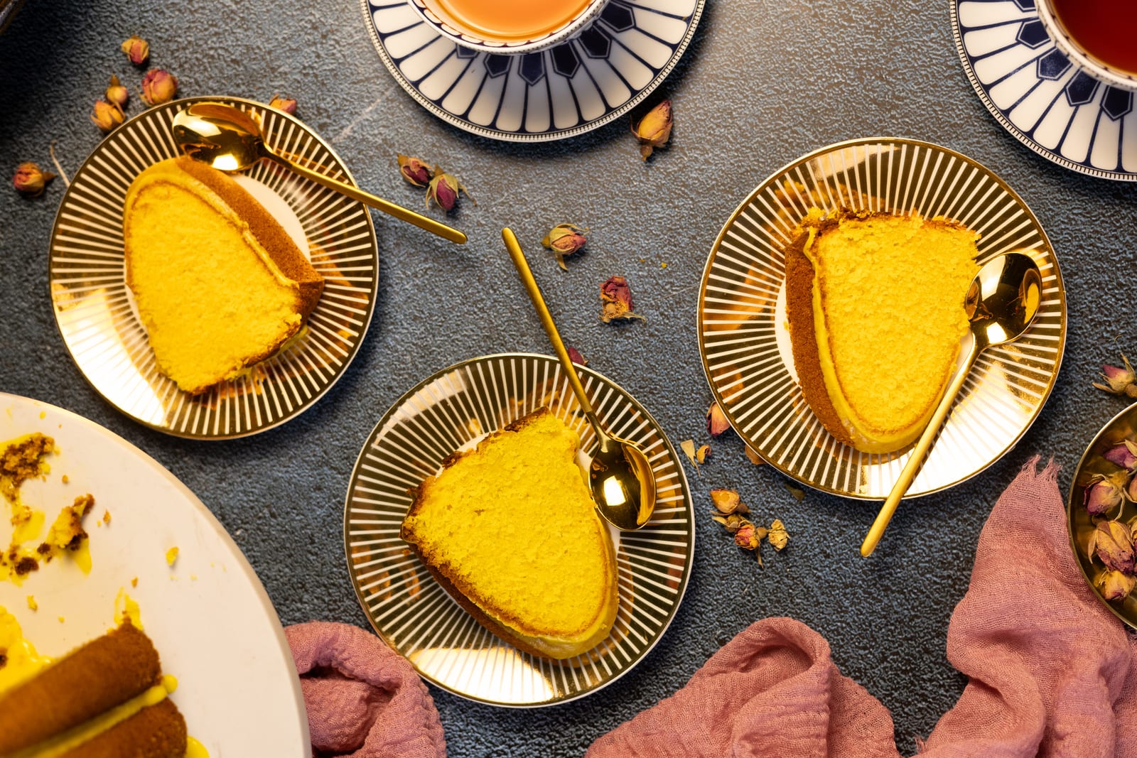 Slices of saffron milk cake on gold-striped plates with gold spoons beside the cut cake, dried rosebuds around, from above