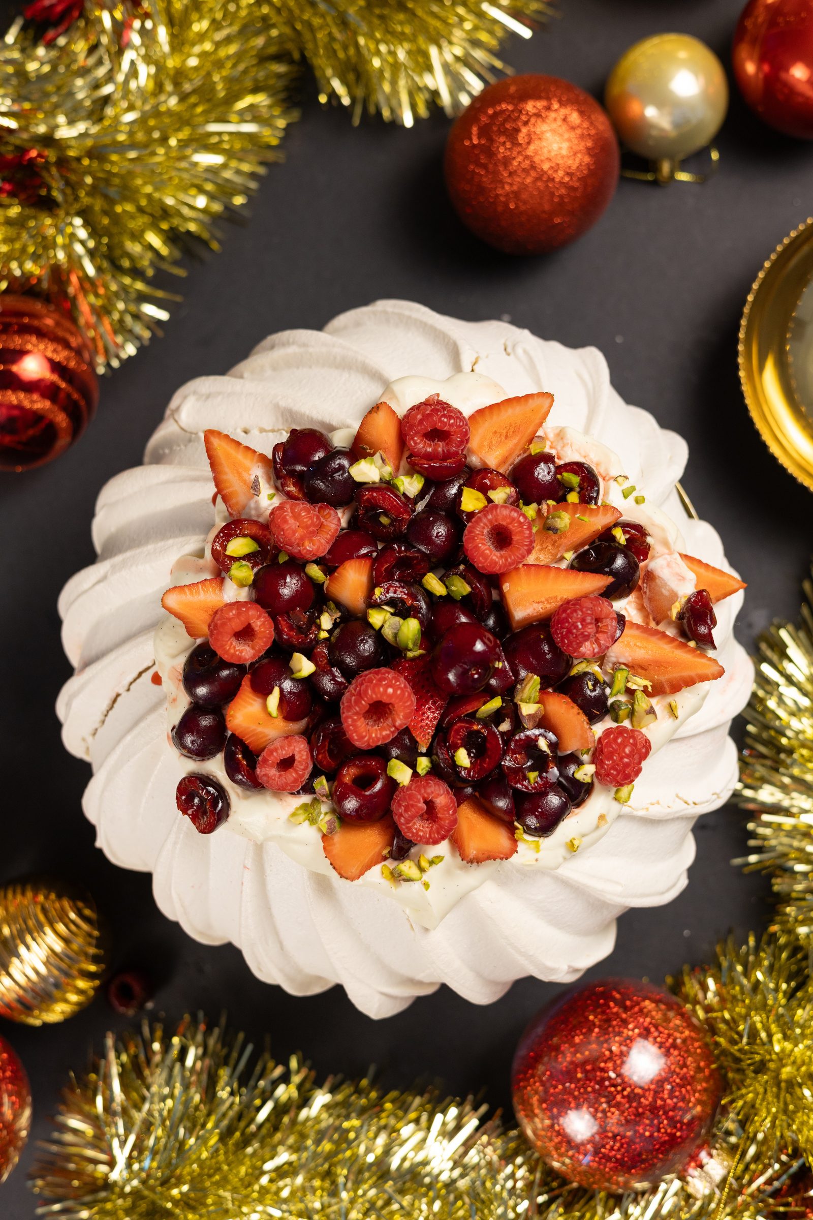 A very berry pavlova filled with cherries, raspberries and pistachios, surrounded by baubles and tinsel, from above