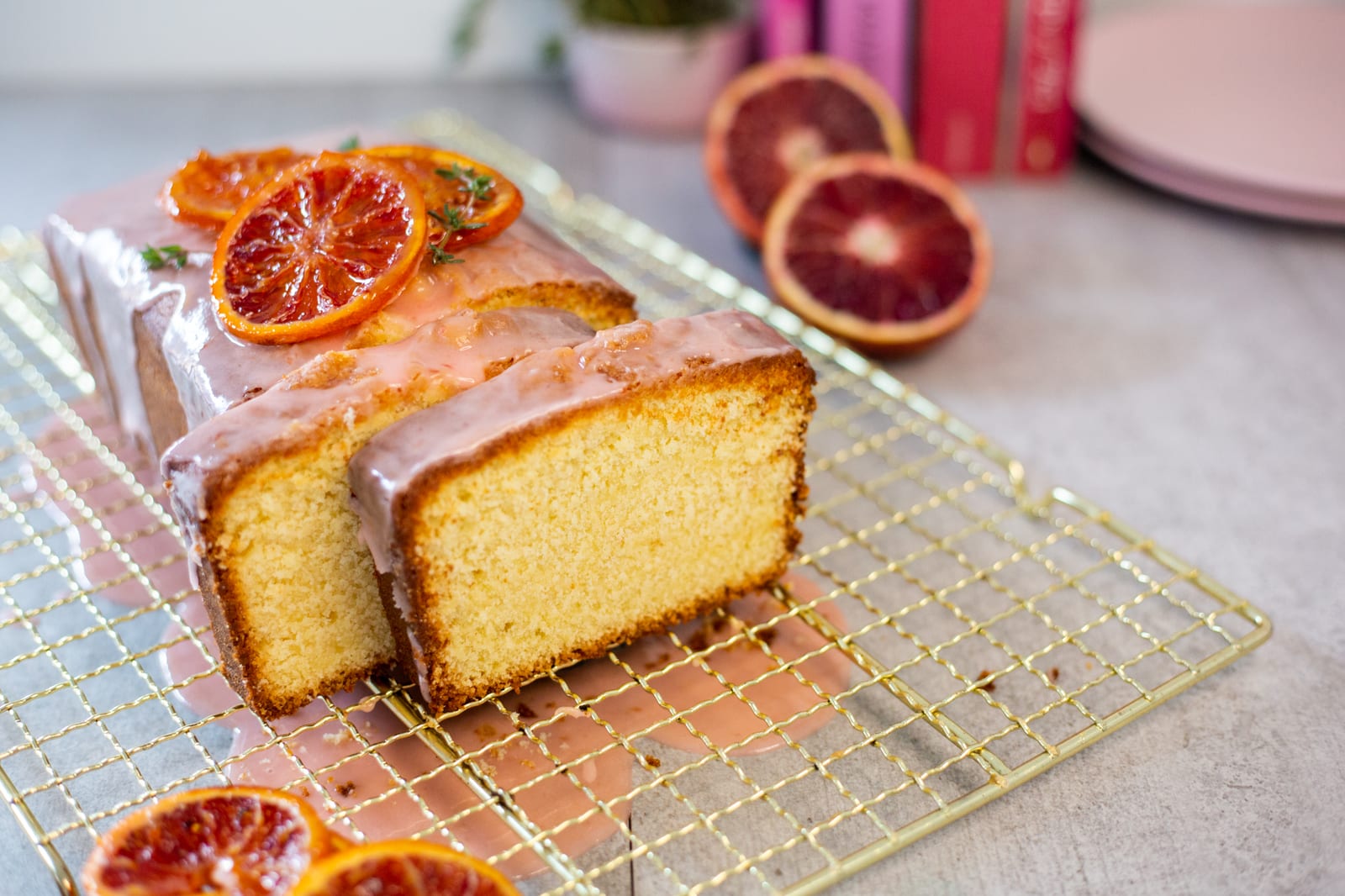 A sliced cream cheese pound cake with blood orange glaze and candied orange slices on a gold rack, pink books behind