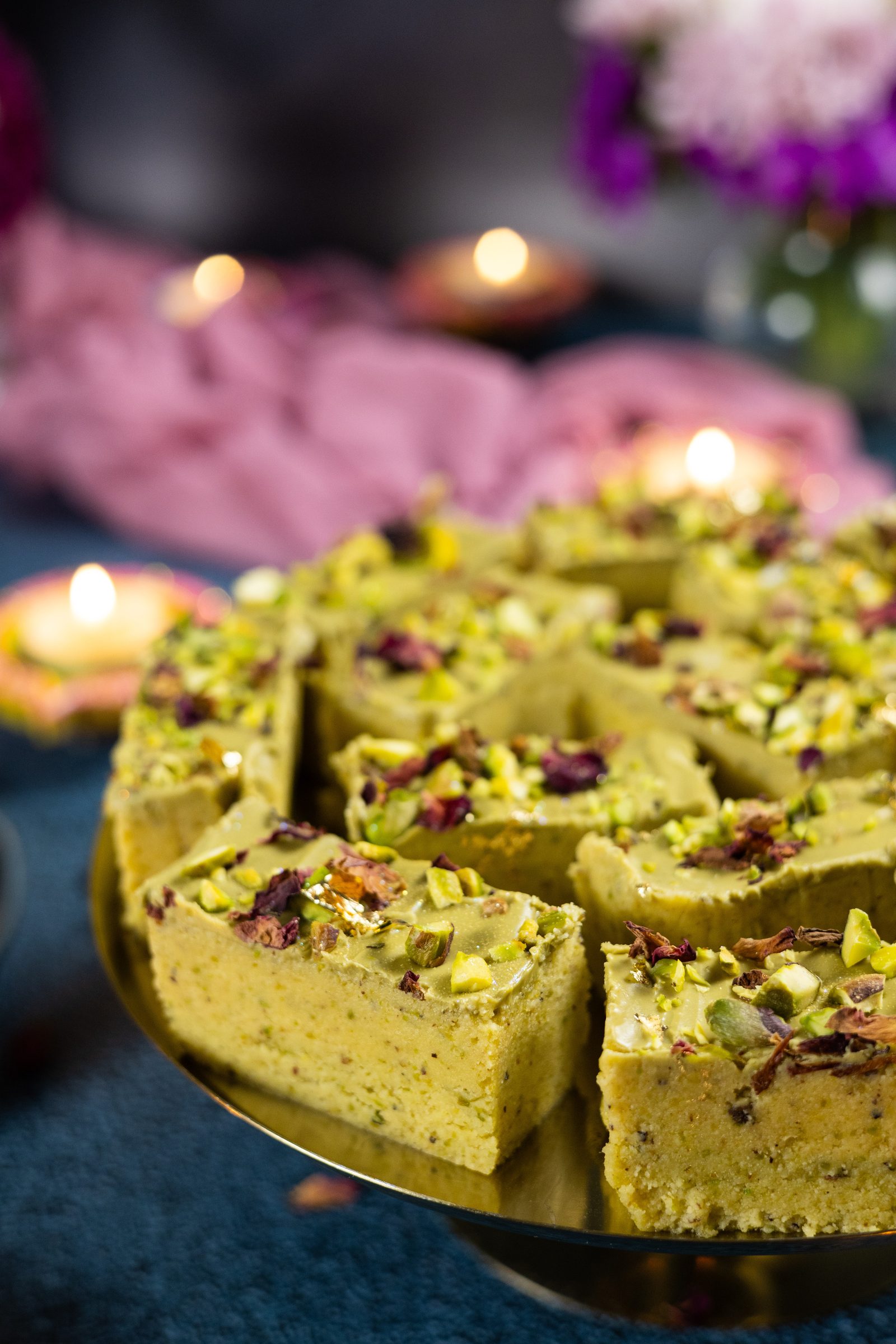 Pistachio burfee on a cake stand with Diwali candles glowing behind, from the side