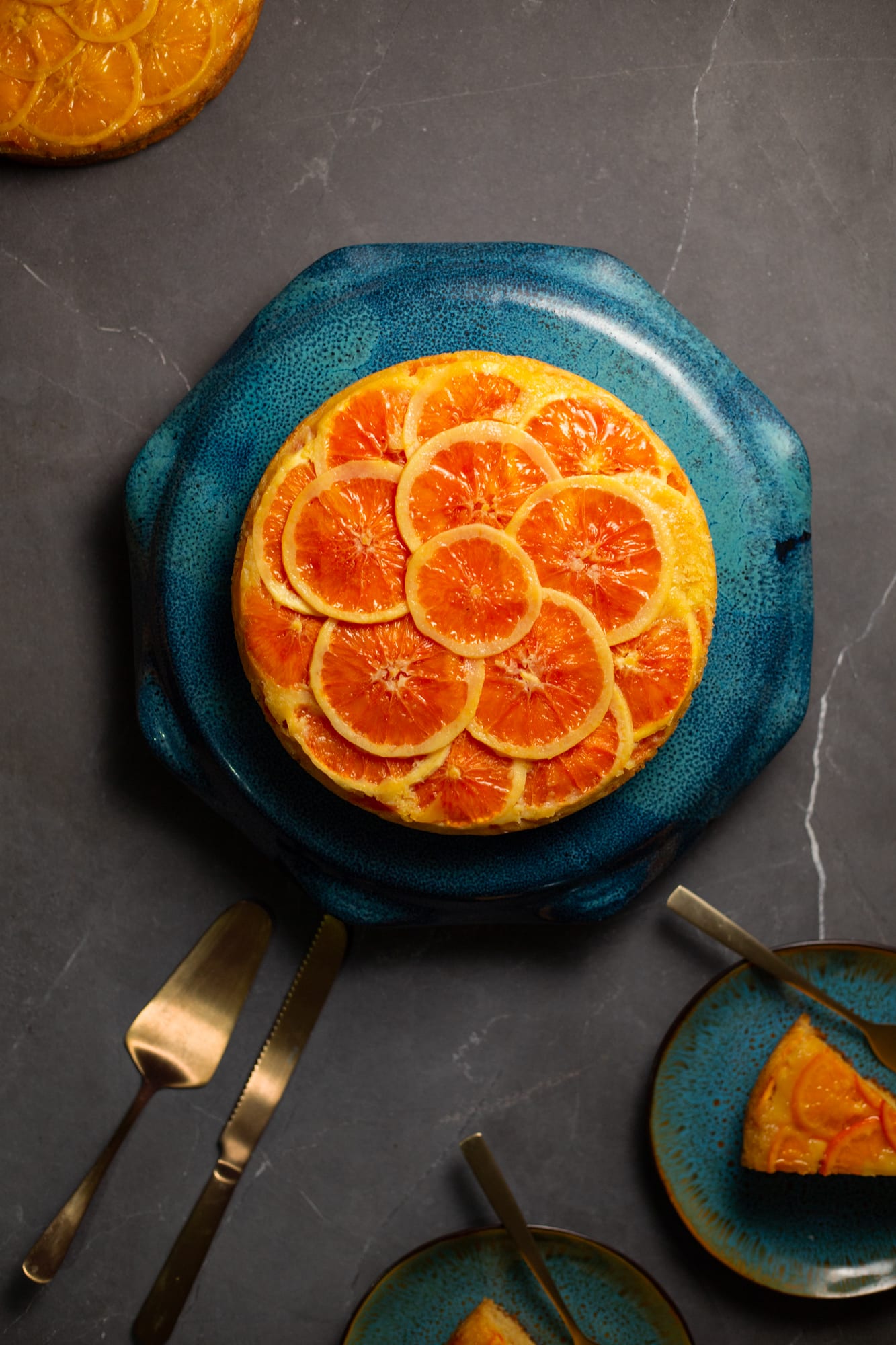 A whole orange upside down cake on a teal plate with a gold cake server and plates of slices, from above
