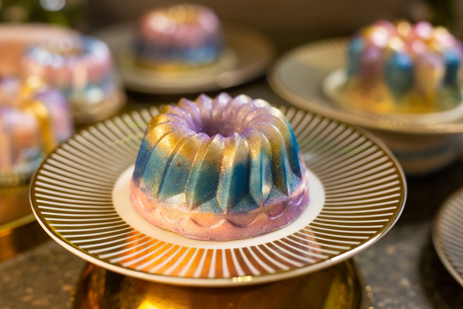 Close-up of a Dubai pistachio burfee bundt on a striped stand, its lustre-dust colours catching the light