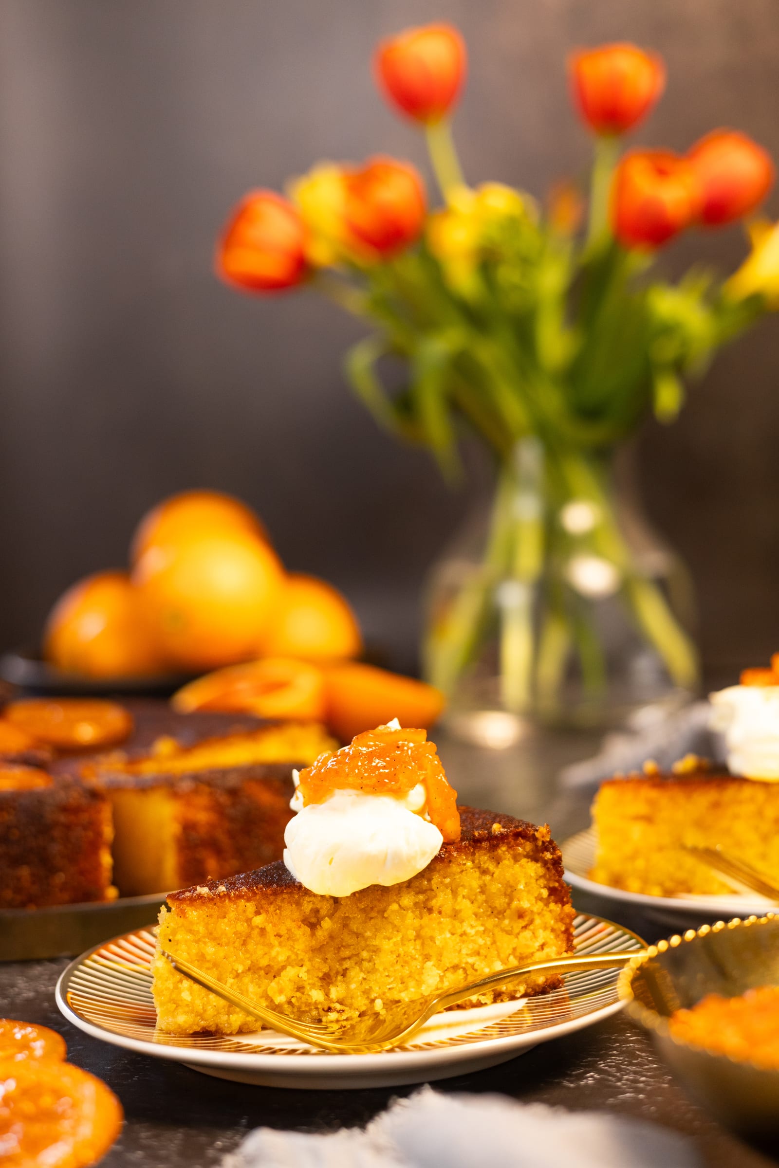 A slice of polenta cake with cream, in front of the cake and a vase of tulips