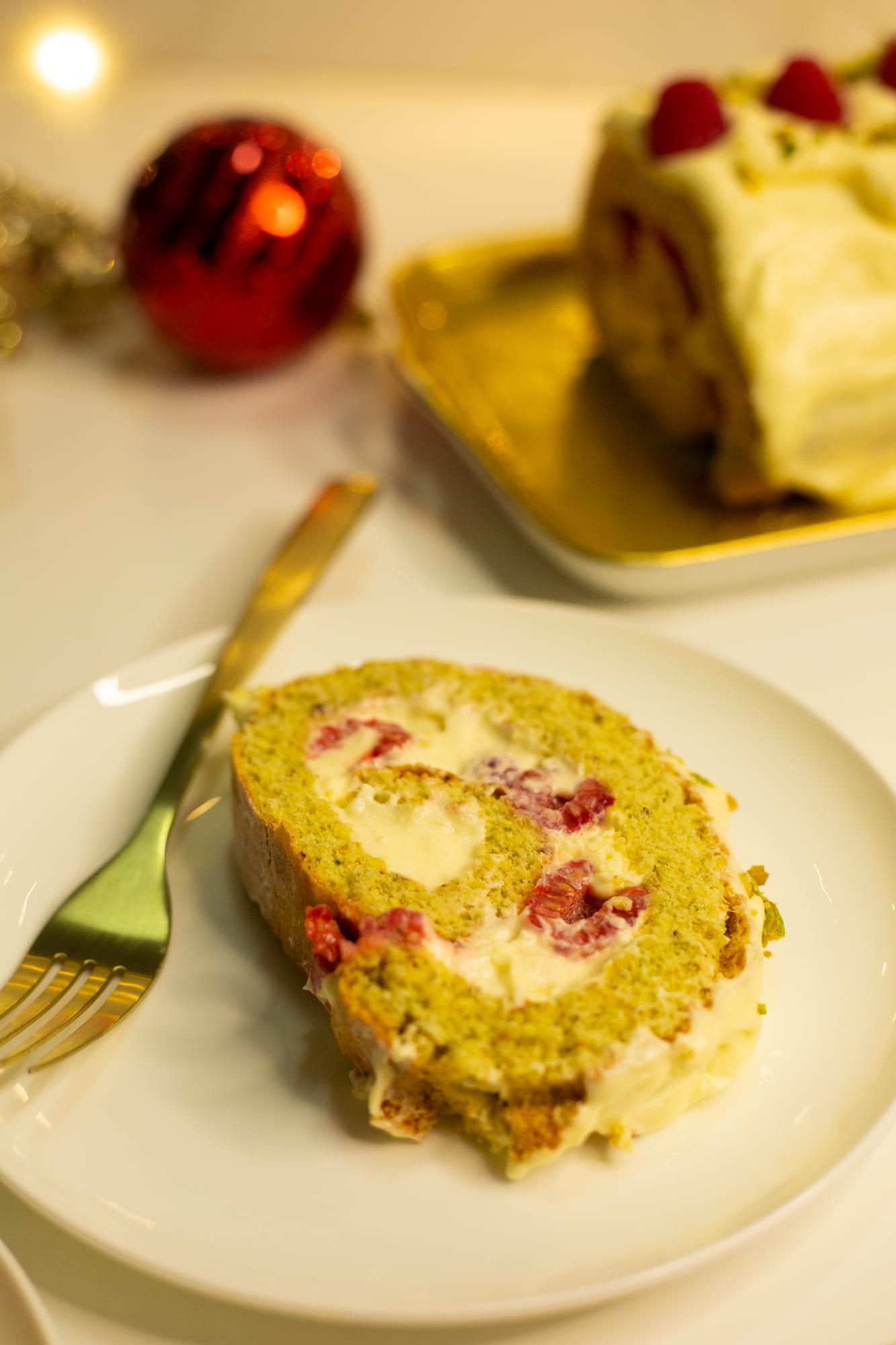 A slice of raspberry and pistachio roulade on a plate with a fork, the roulade behind