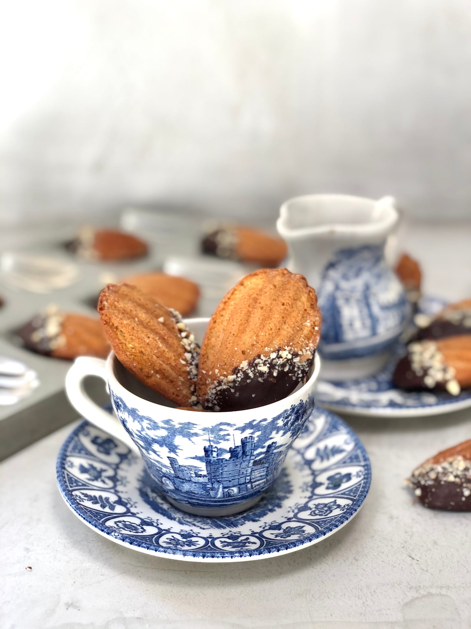 A teacup heaped with brown butter and almond madeleines, a blue and white jug behind