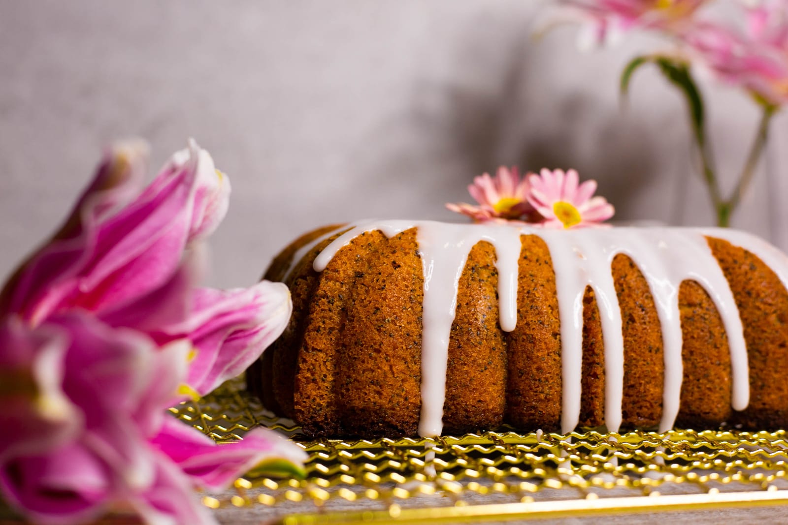 A glazed gluten-free lemon and poppyseed loaf with a pink daisy on a gold rack, pink lilies in front