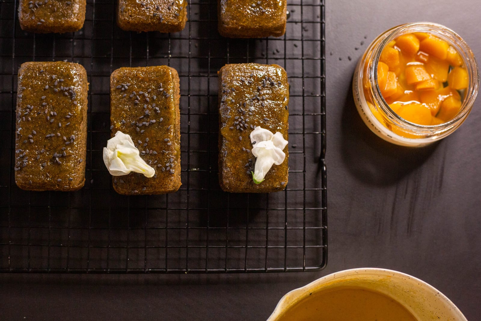 Two rows of black sesame and ginger financiers with cream on a wire rack beside a jar of ginger, from above