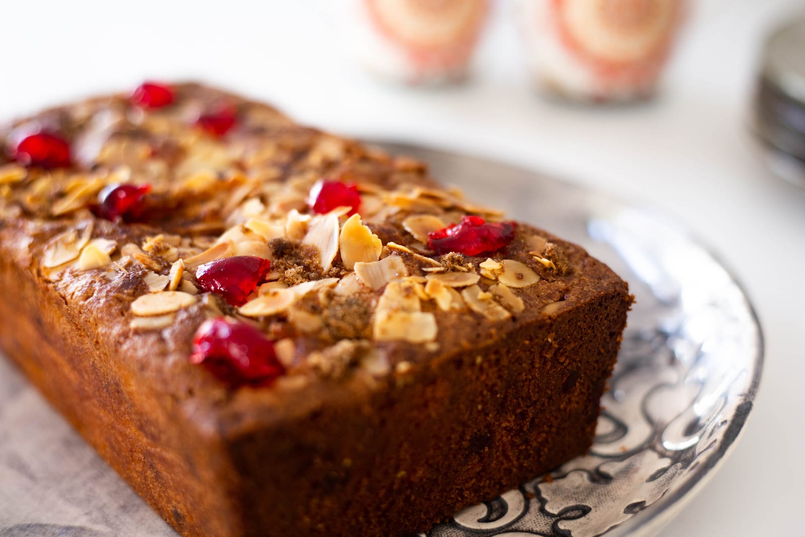 A gluten-free cherry and almond loaf on a patterned plate in bright daylight, mugs blurred behind