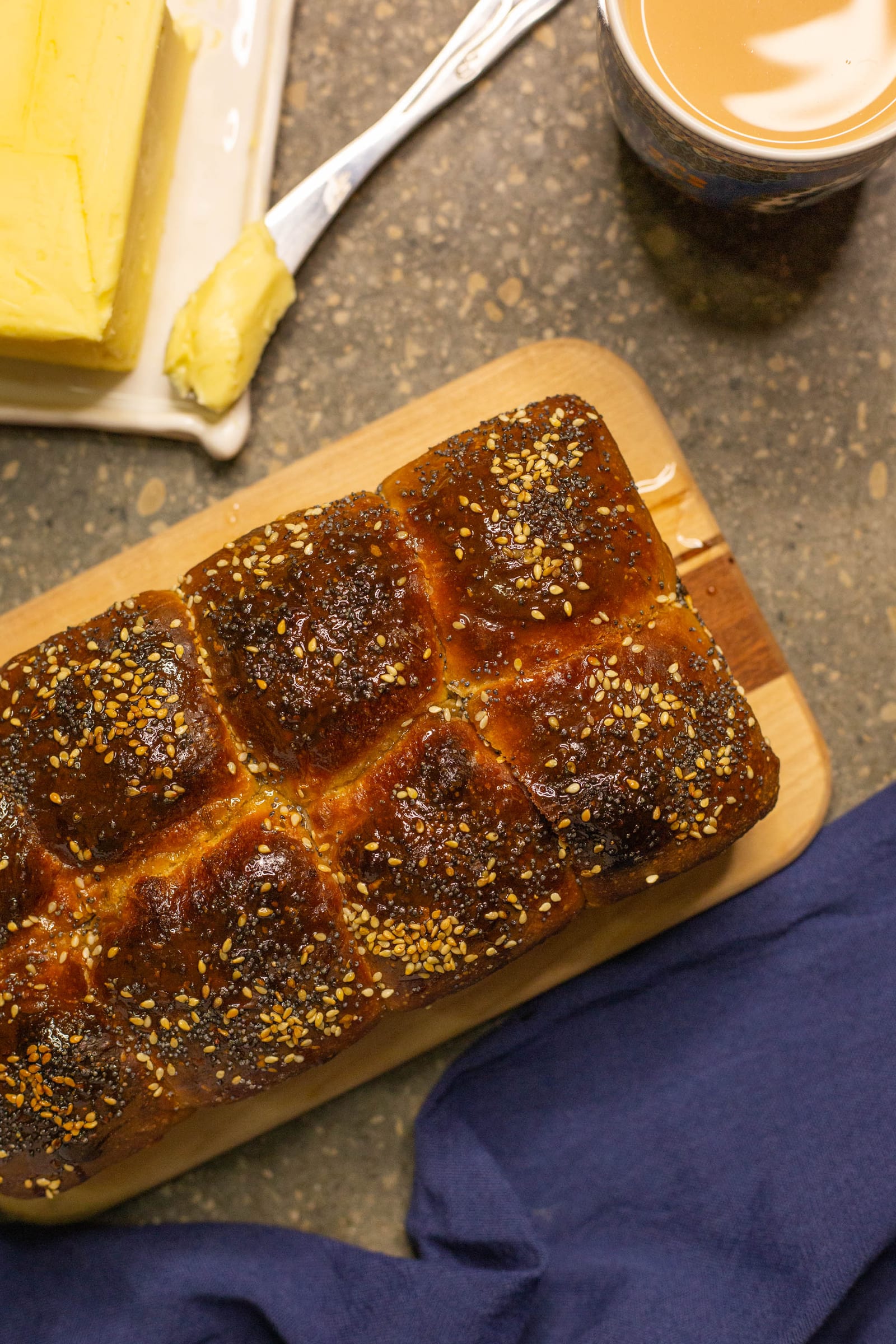 A mosbolletjies loaf topped with poppy and sesame seeds, beside butter and tea