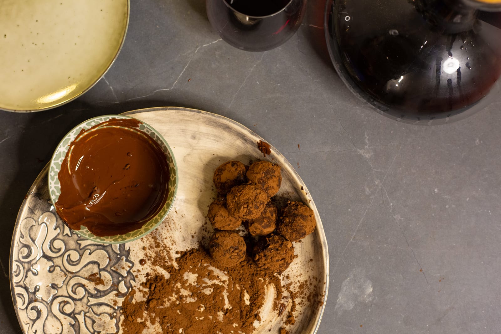 Rum and raisin truffles on a plate with cocoa powder and a bowl of melted chocolate, wine glasses beside them, from above
