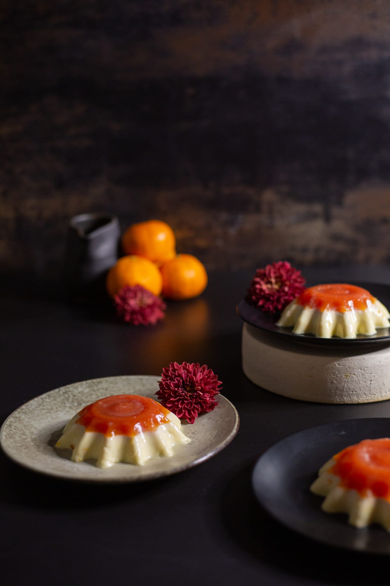 Two vanilla yoghurt panna cottas with blood orange jelly on beige and black plates, beside oranges and a jug
