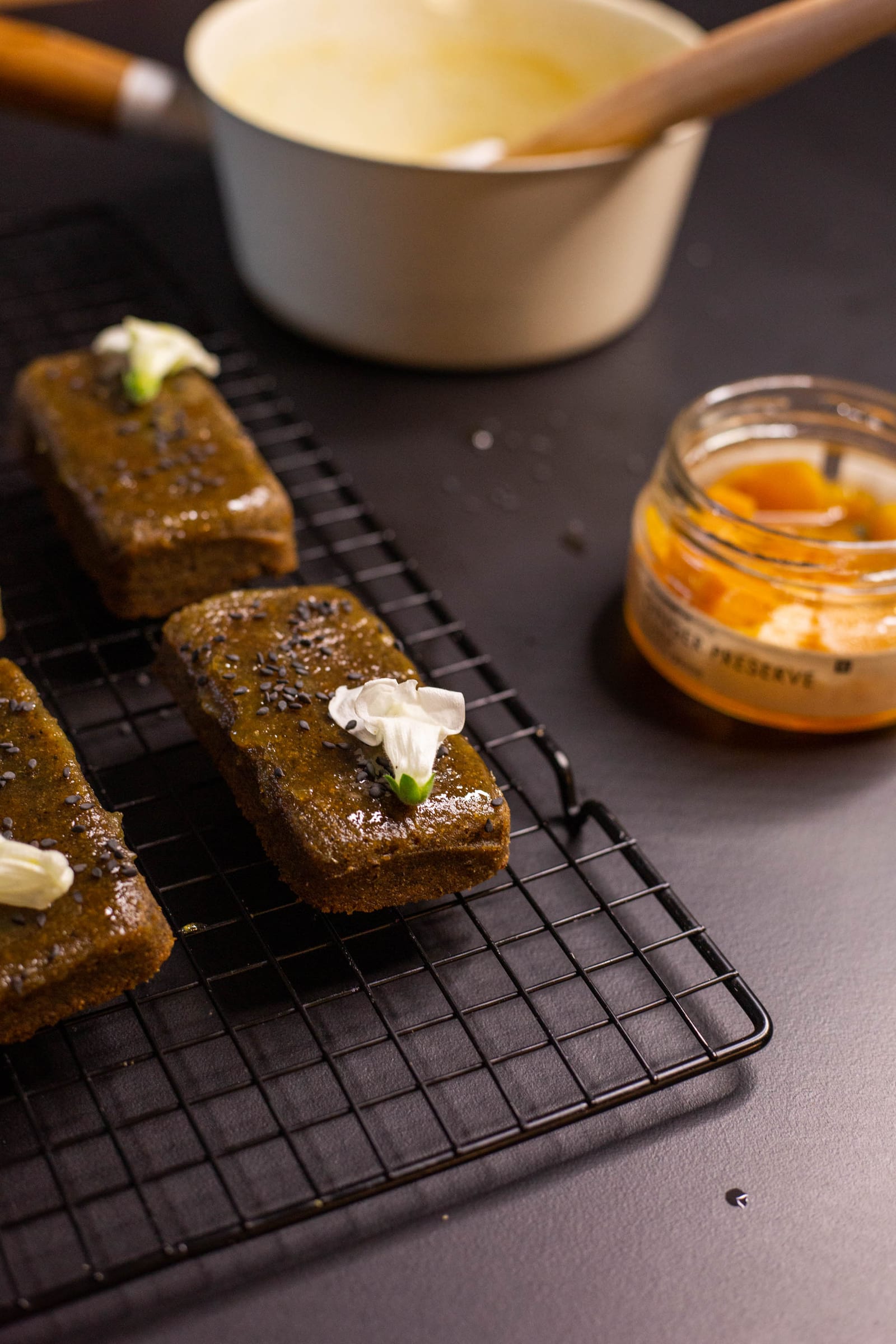 Black sesame and ginger financiers on a wire rack with a bowl of cream and a jar of ginger behind