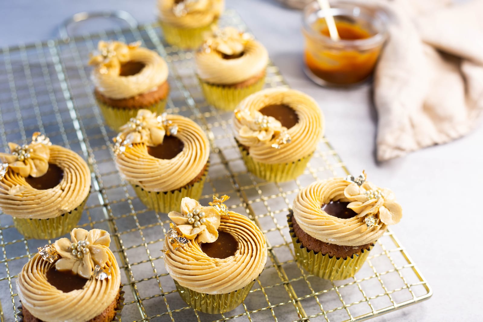Salted caramel cupcakes with gold sugar flowers on a gold wire rack, beside a jar of salted caramel sauce