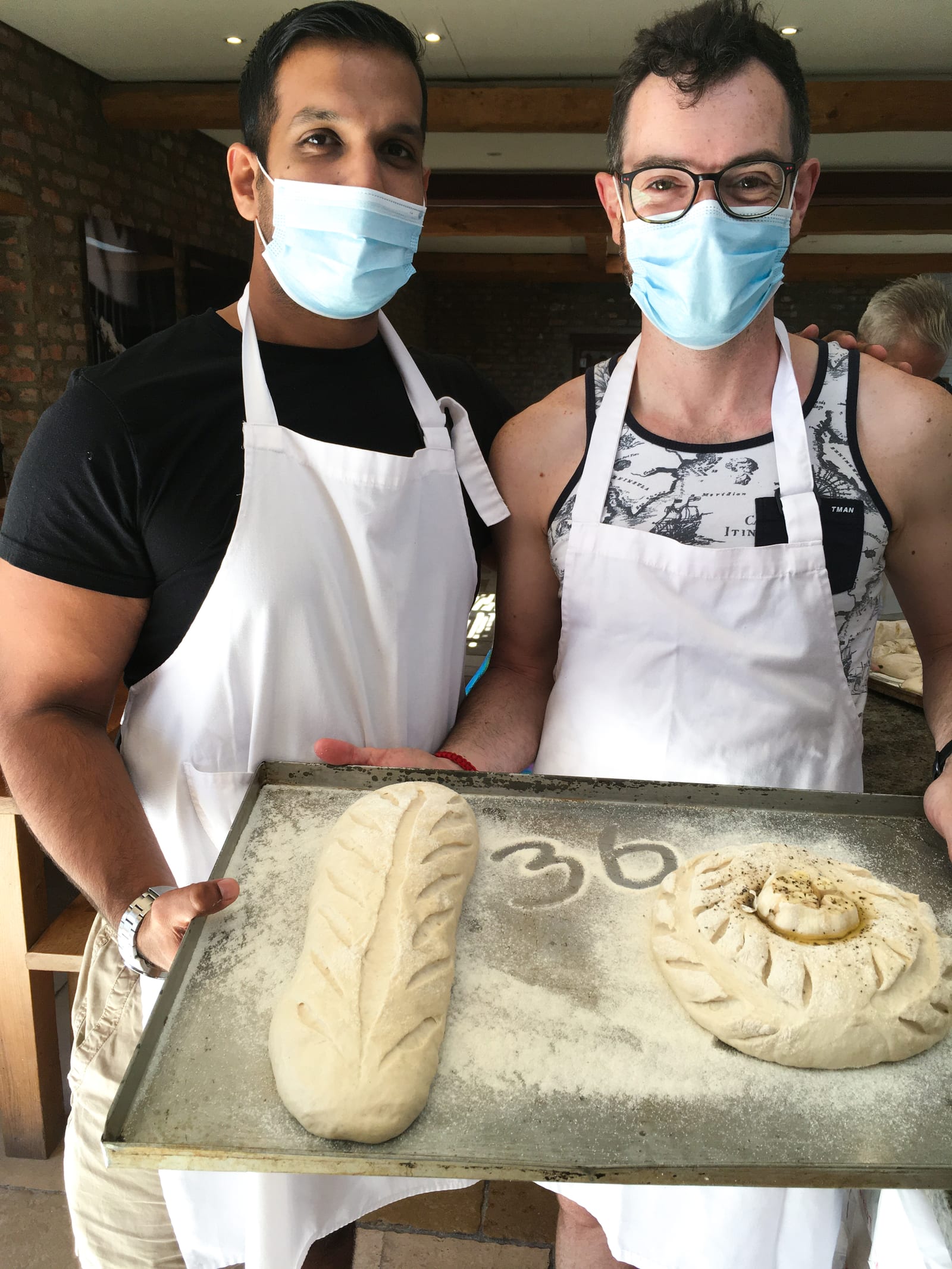 Suvar (left) and Linsen (right) in aprons at the Babylonstoren bakery, holding a floured tray of their scored sourdough: a long loaf and a round garlic bread