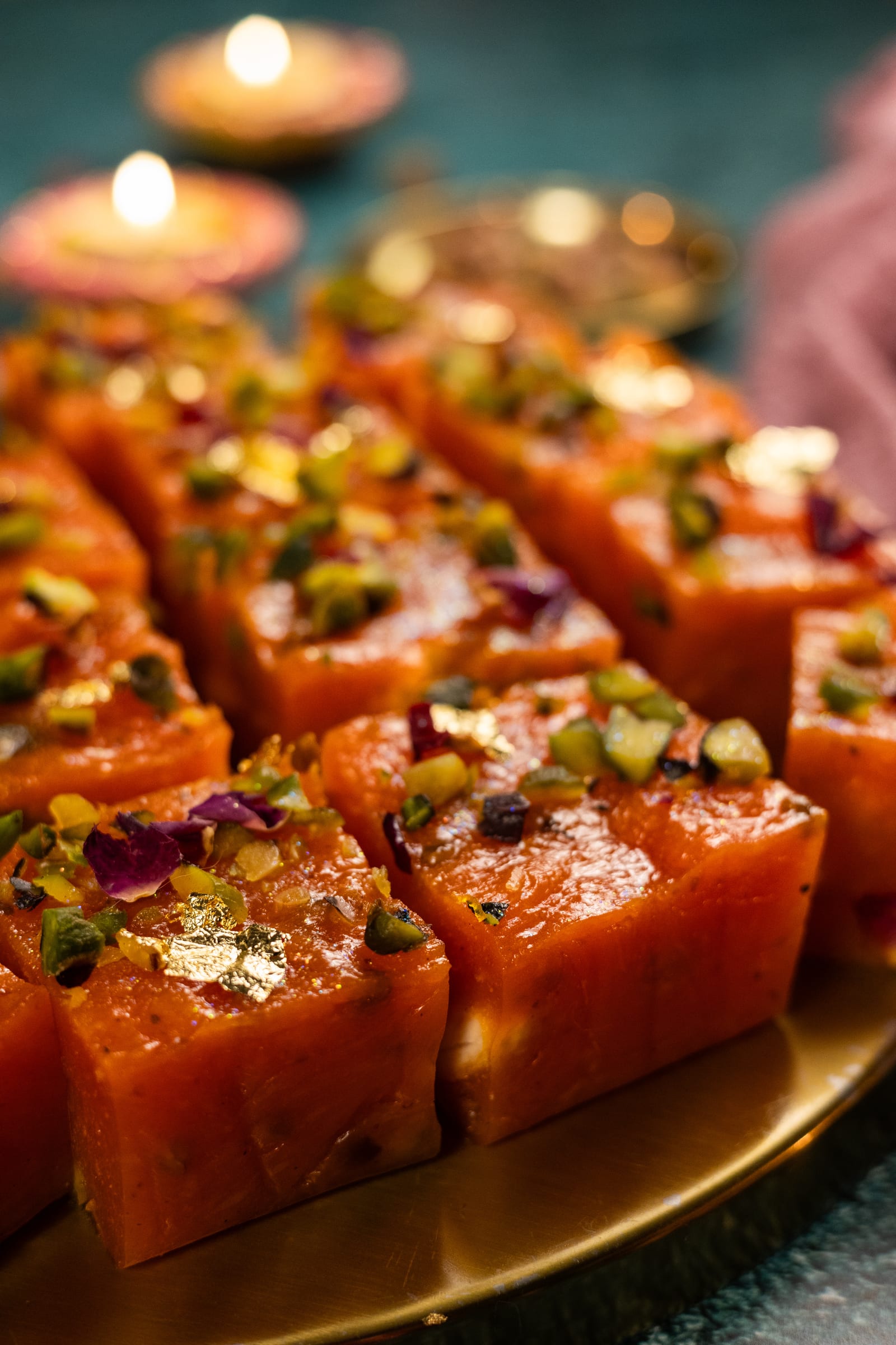 Glossy blocks of halwa at eye level, with lit diyas glowing behind