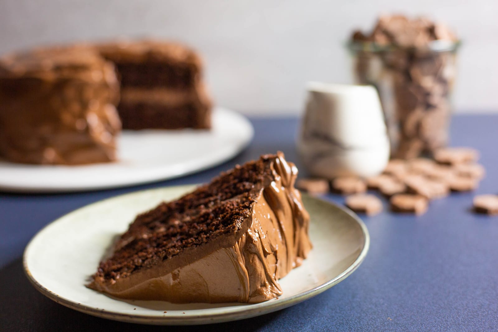 A slice of chocolate fudge cake on a plate in front of the cut cake, a white jug behind