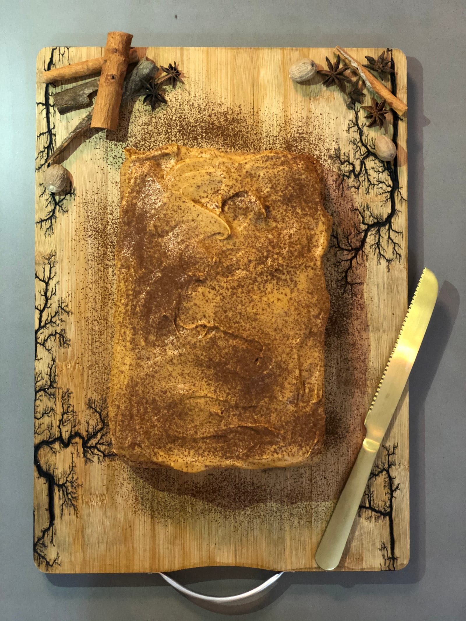 A whole masala tres leches cake dusted with spice on a patterned board with a gold knife and cinnamon, from above