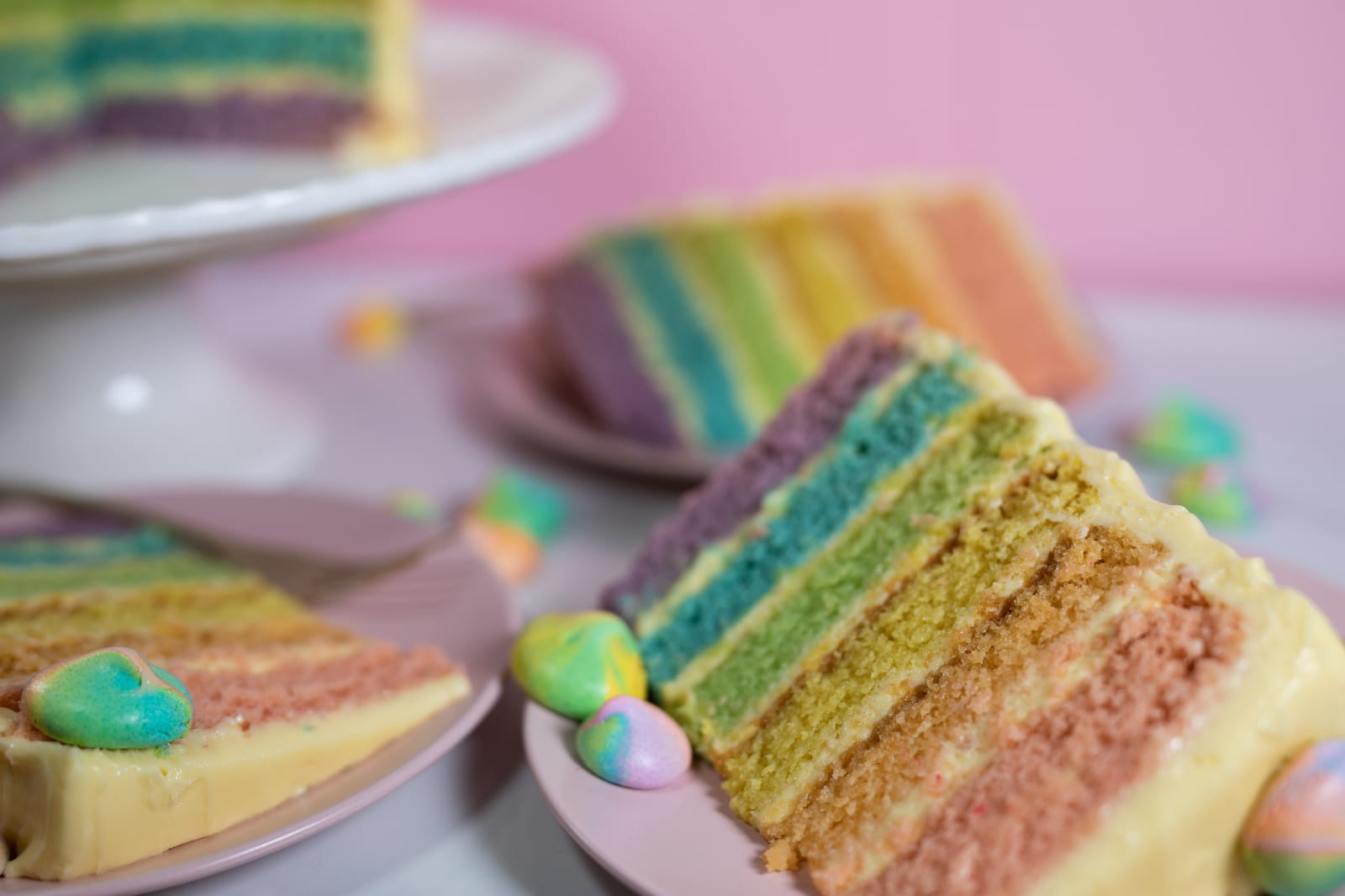 A slice of pastel rainbow cake with meringue kisses in front, the cake stand behind