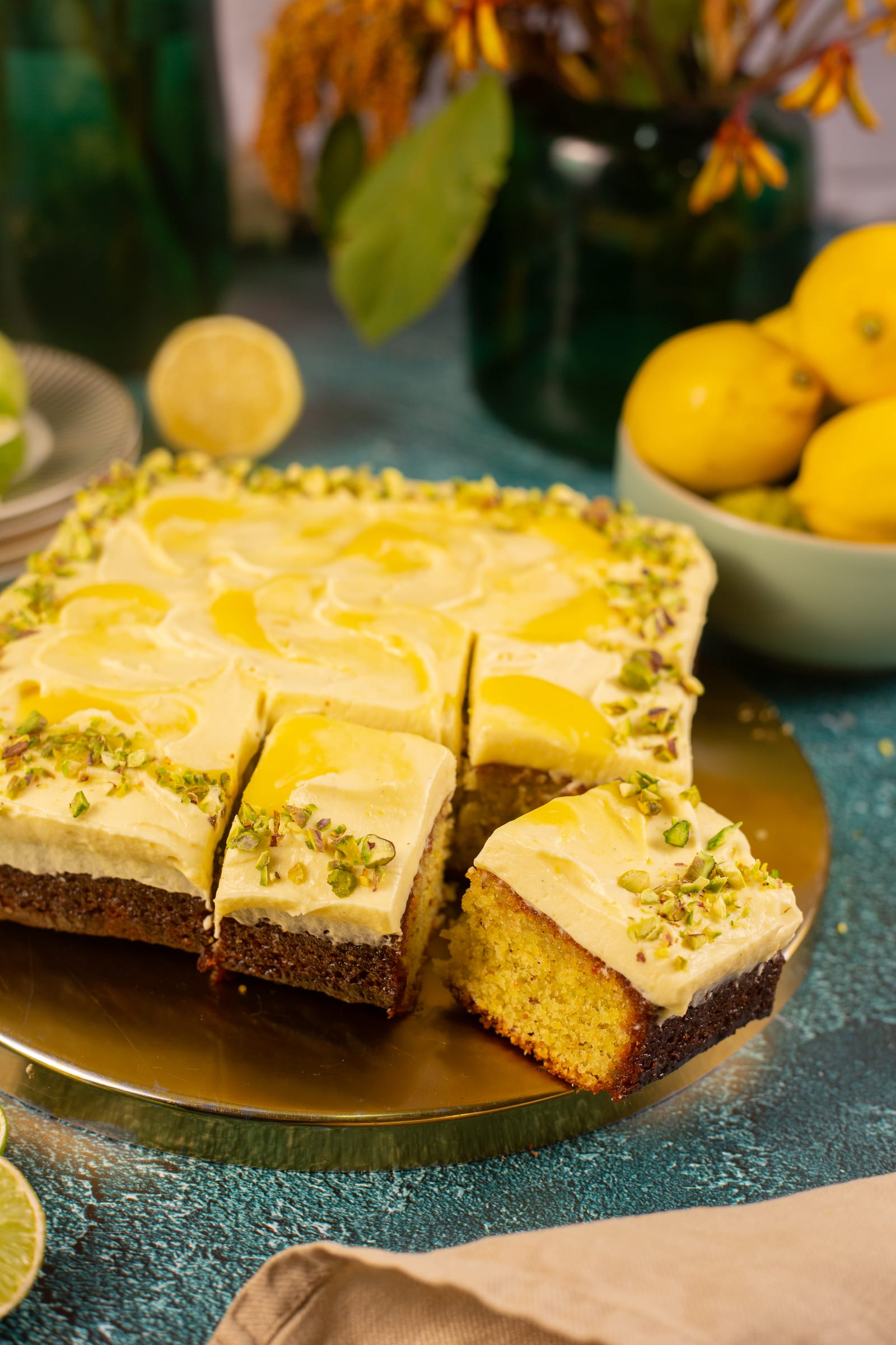 The pistachio and lemon traybake cut into squares, with flowers and lemons behind
