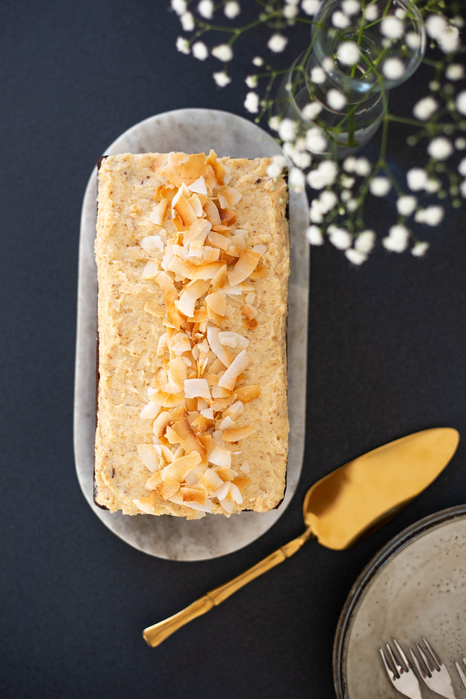 A gluten-free coconut loaf topped with toasted coconut flakes on a marble board, baby's breath above, from above
