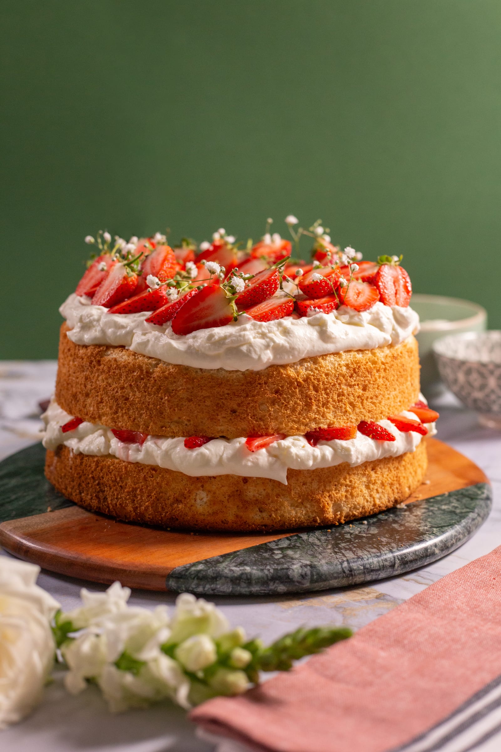 A strawberry and cream angel food cake on a marble and wood board, white flowers beside it