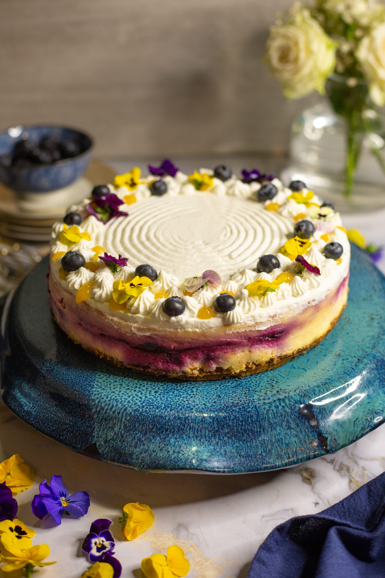 A whole lemon and blueberry cheesecake on a teal stand, topped with whipped cream, blueberries and edible flowers