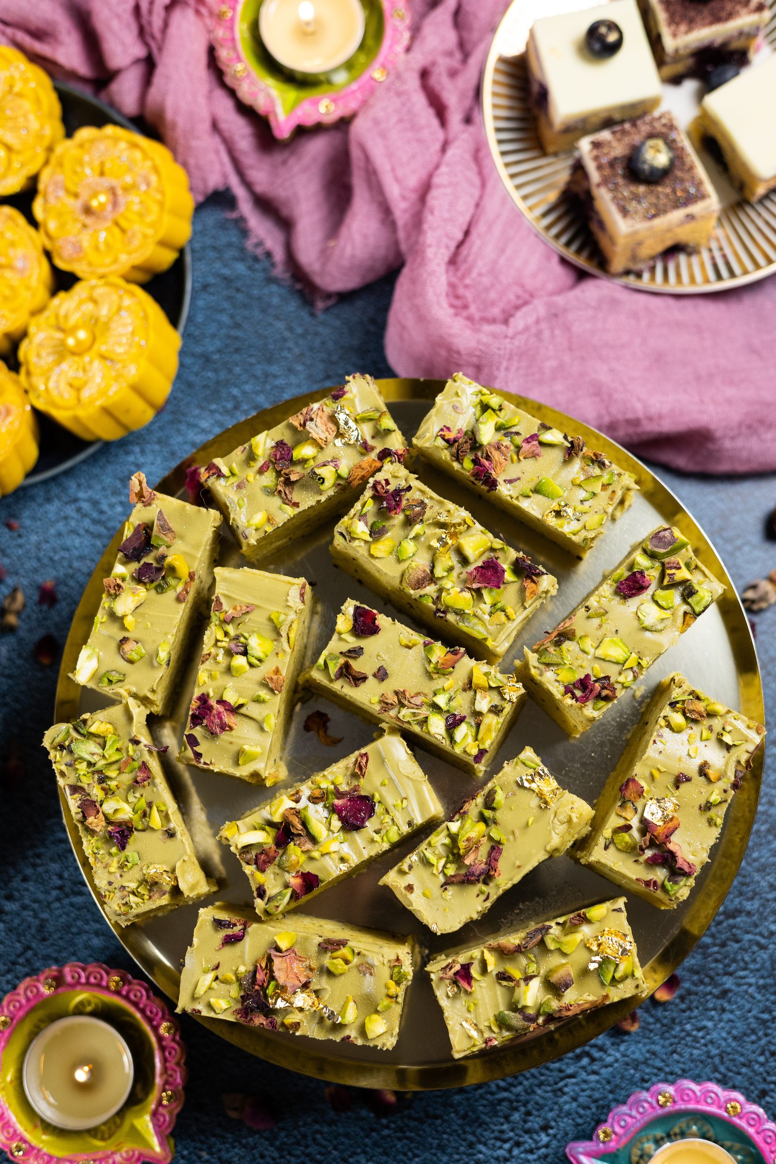 A gold plate of pistachio burfee with mango burfee and blueberry ripple burfee beside it, from above