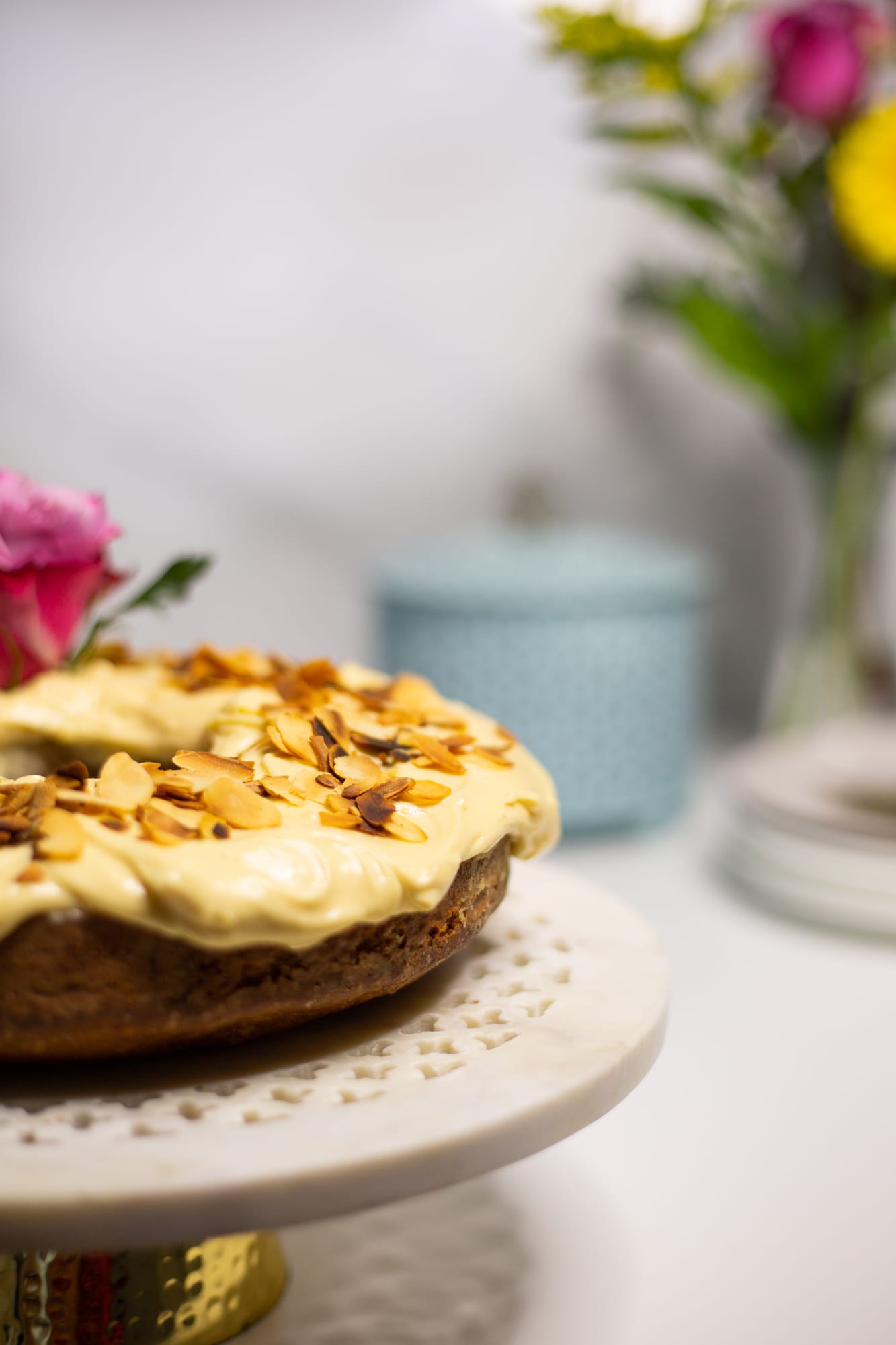 A citrus pound cake with brown sugar cream cheese icing on a white lace-edged cake stand, flowers behind