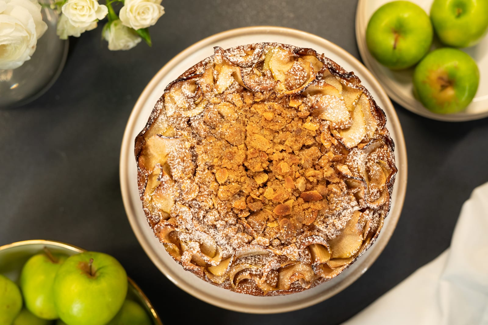 An apple crumble cake from above, its crumble centre ringed with sliced apples, green apples around it