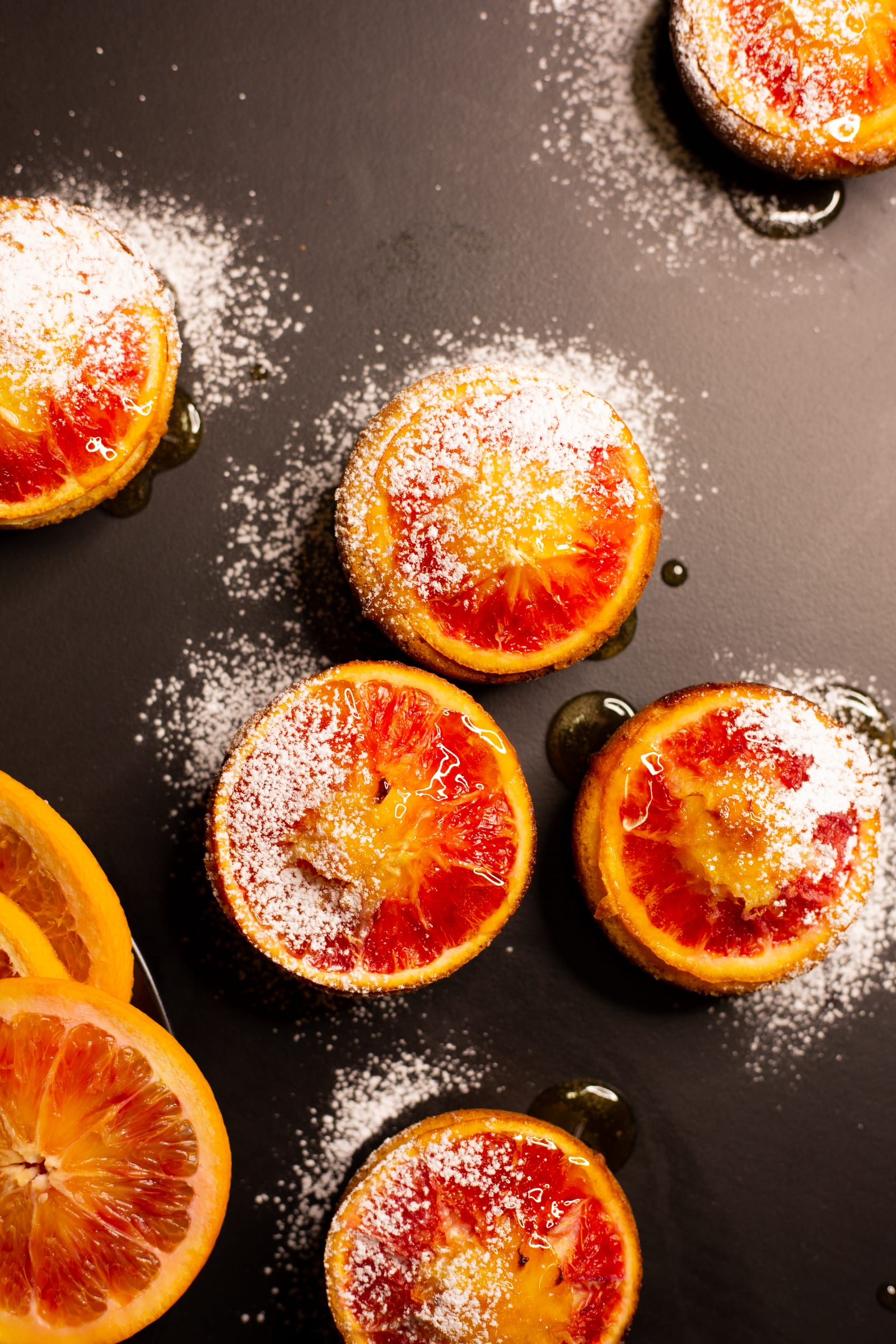Almond, semolina and blood orange syrup cakes dusted with icing sugar beside blood orange halves, from above