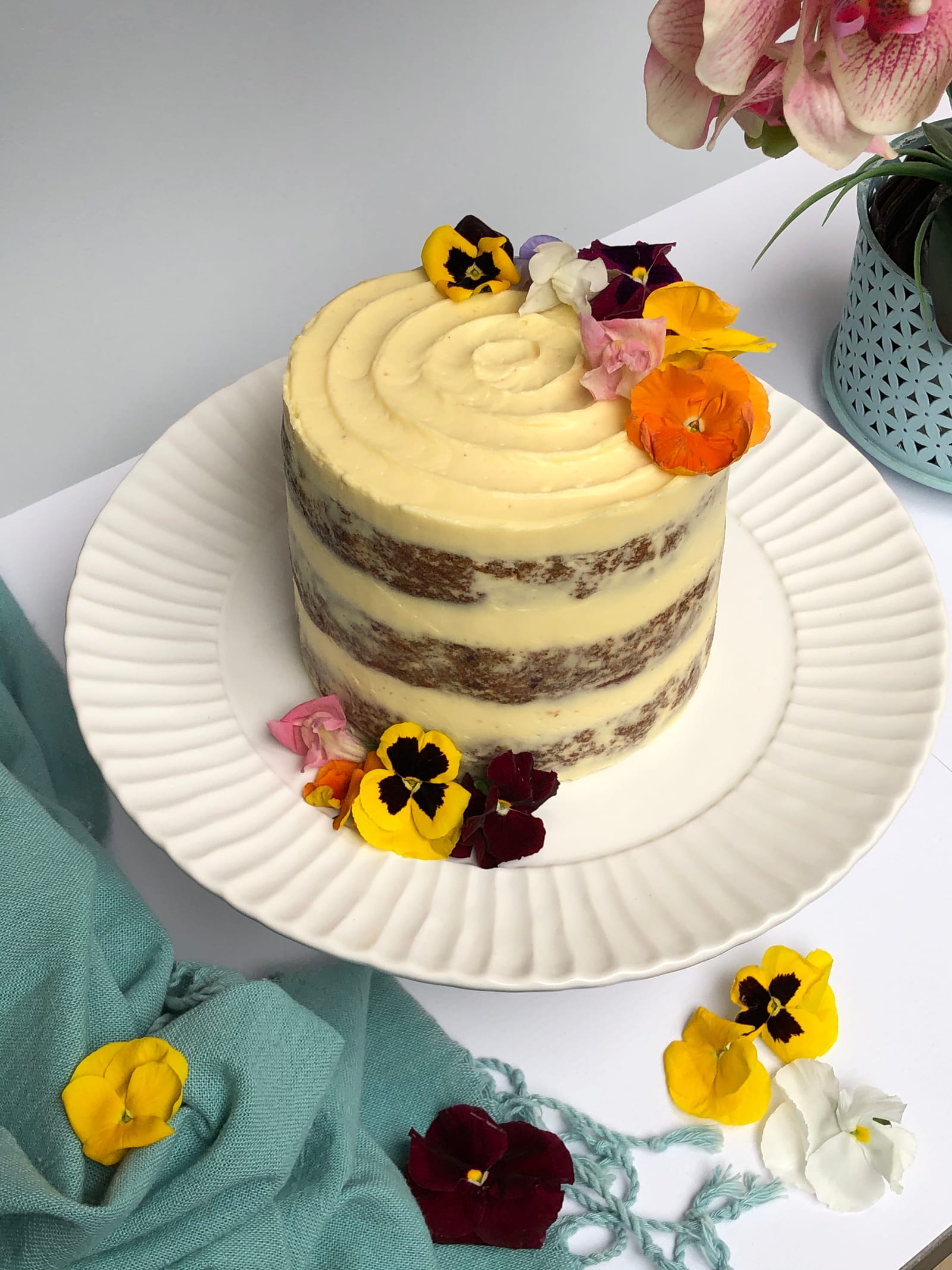 A naked layer carrot cake topped with flowers on a teal cloth with pansies scattered around
